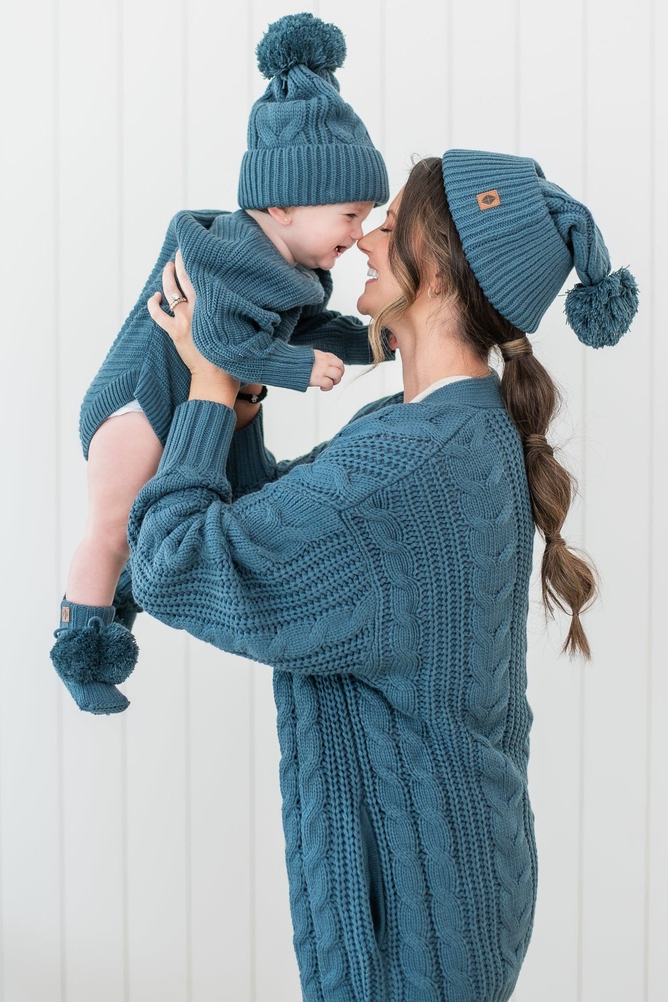 Female model holding her son up to her face with their noses touching. Mother wearing the Cable Knit Adult Cardigan in Atlantic and Cable Knit Pom beanie with her child matching in an Atlantic chunky knit romper, knit booties and cable knit pom beanie
