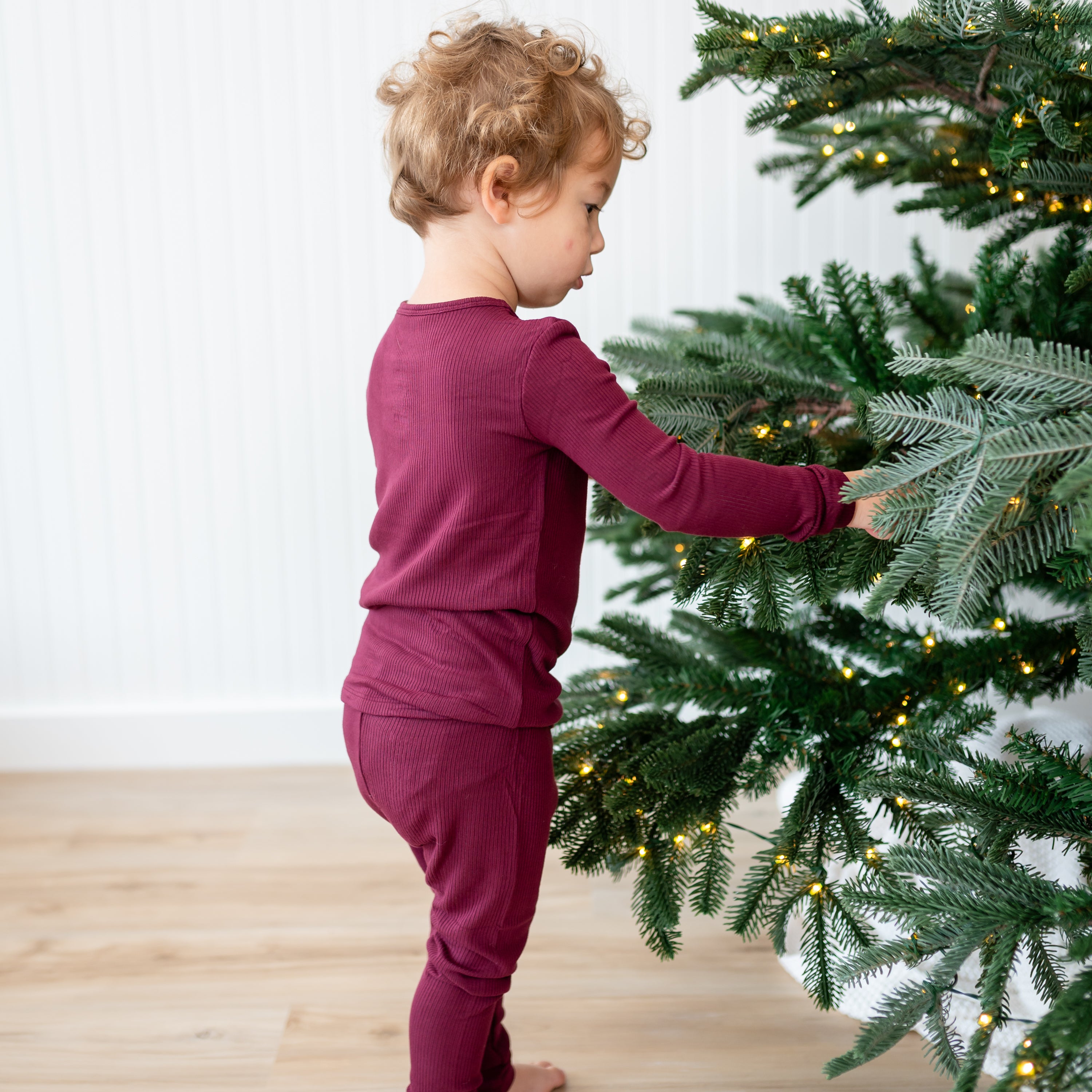 Young toddler touching the lights on a lit Christmas tree wearing the Ribbed Henley Set in Burgundy
