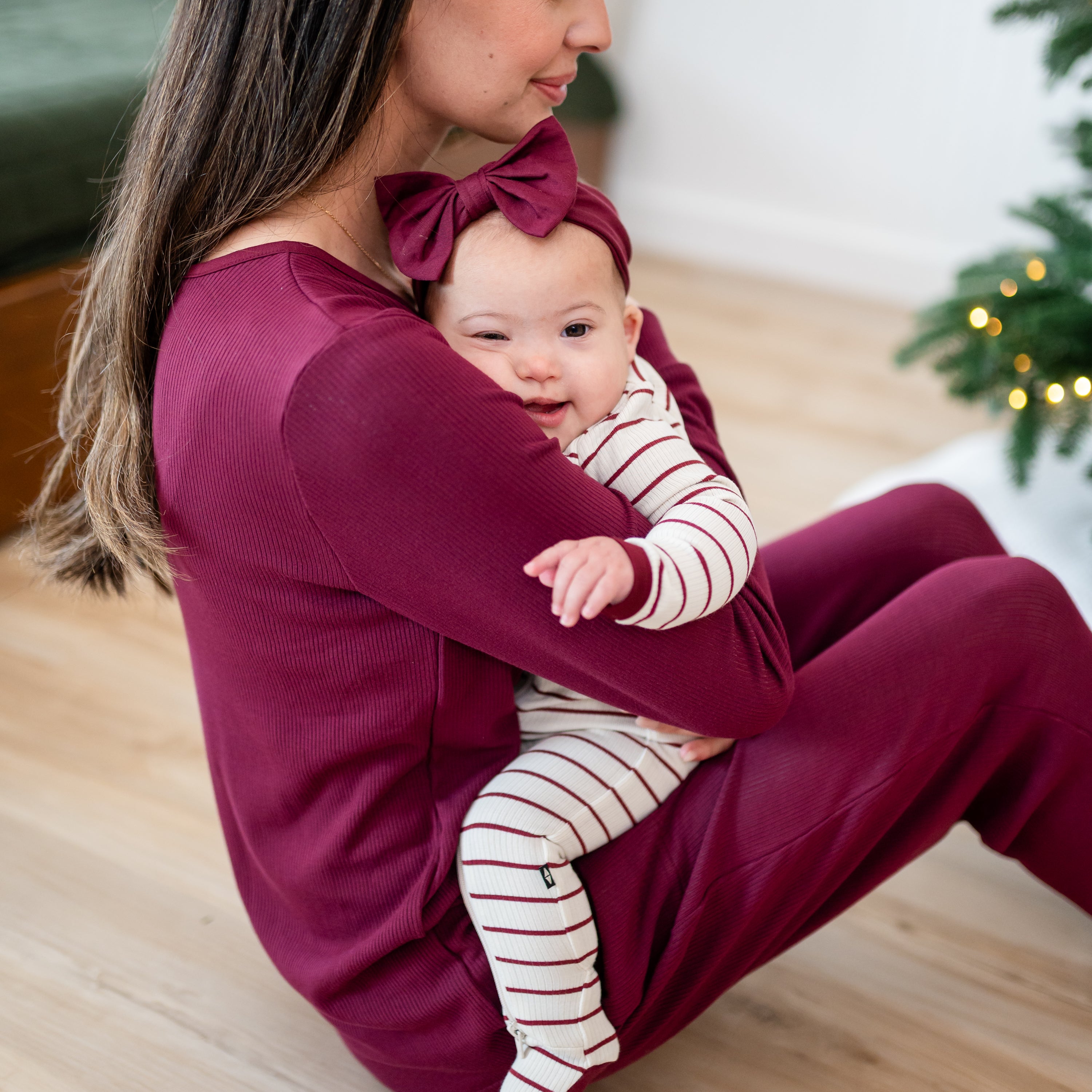 Mother sitting on the floor wearing the Women's Ribbed Henley set in Burgundy holding her daughter who is wearing the Ribbed Zipper Footie in Burgundy Stripe
