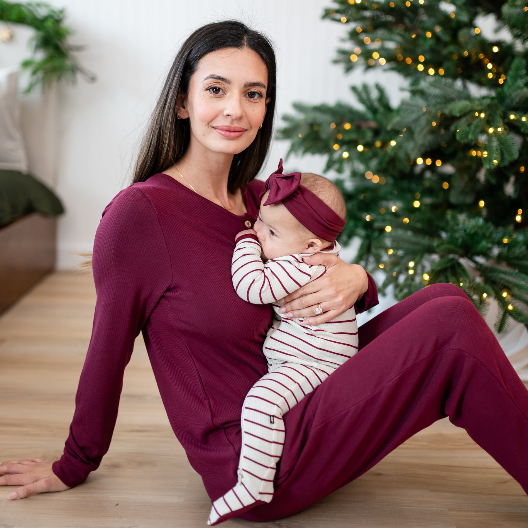 Mother sitting on the floor wearing the Women’s Ribbed Henley Set in Burgundy holding her infant daughter who is wearing the Ribbed Zipper Footie in Burgundy Stripe