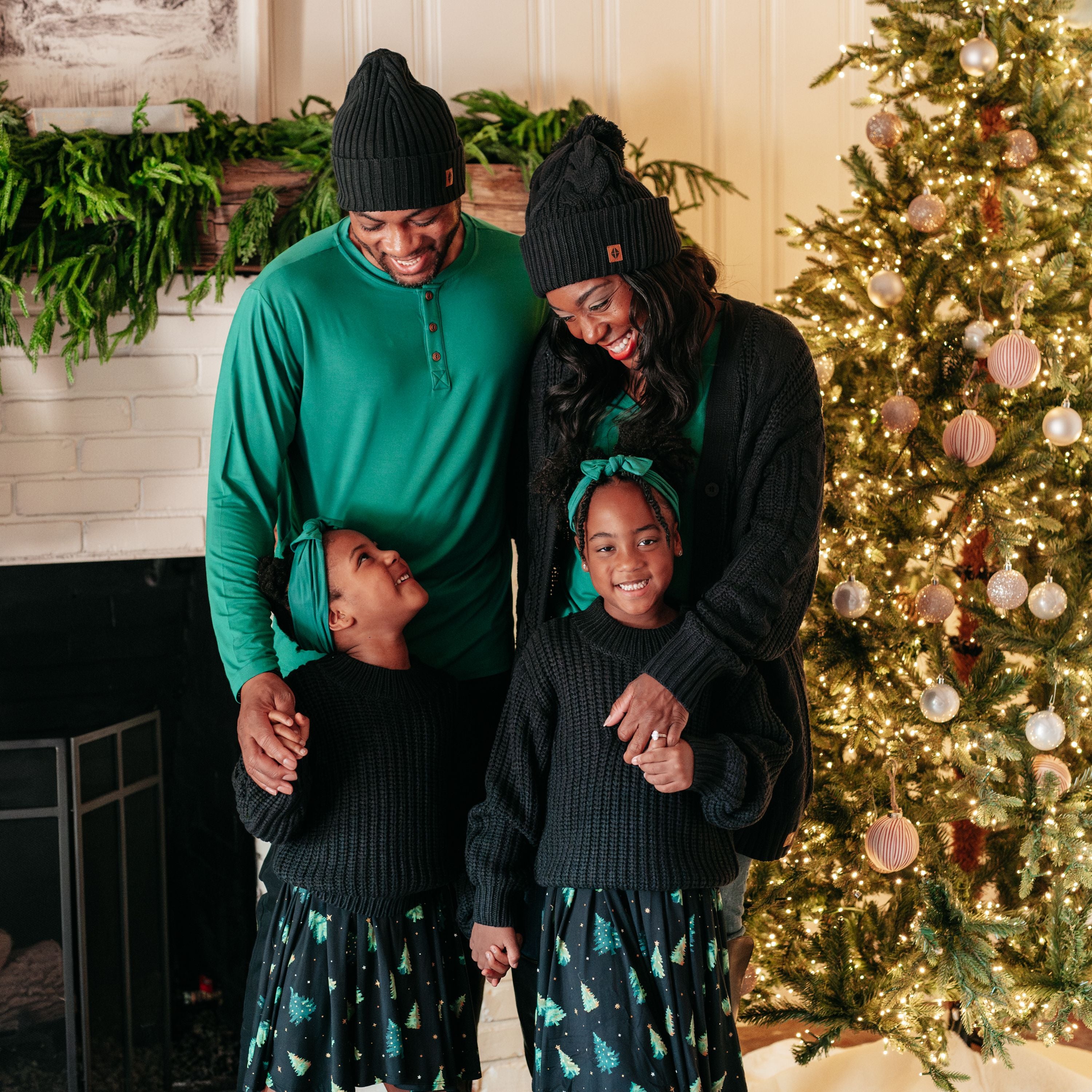 Family of four in coordinating colors standing beside a Decorated Christmas tree. Two daughters wearing the Long Sleeve Twirl Dress in Twinkle Tree and Midnight Chunky knit sweaters overtop. Father and mother wearing Emerald tops and Black beanies