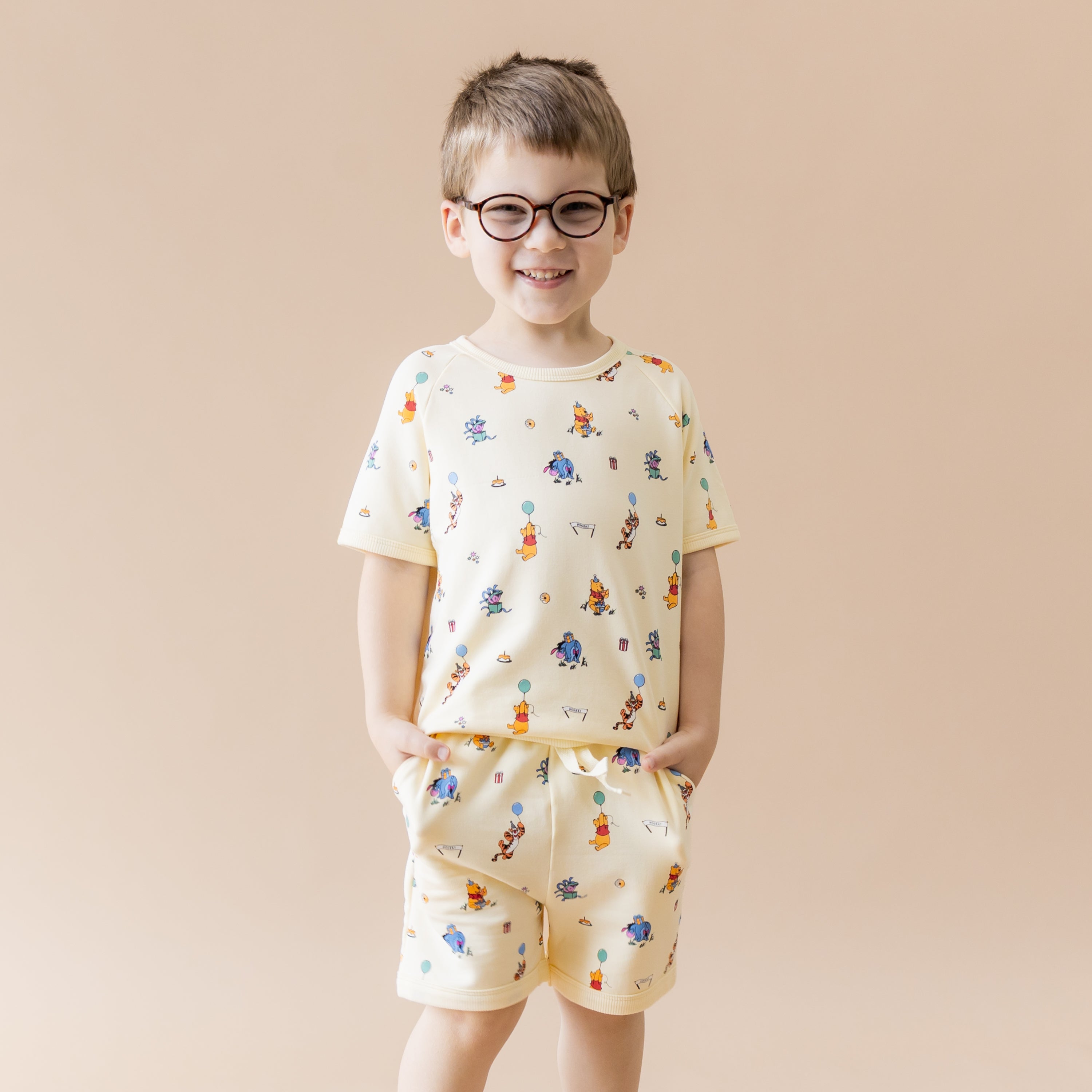 Young boy modeling the Short Sleeve Jogger Set in Winnie the Pooh Celebration with both hands in the pockets of the shorts