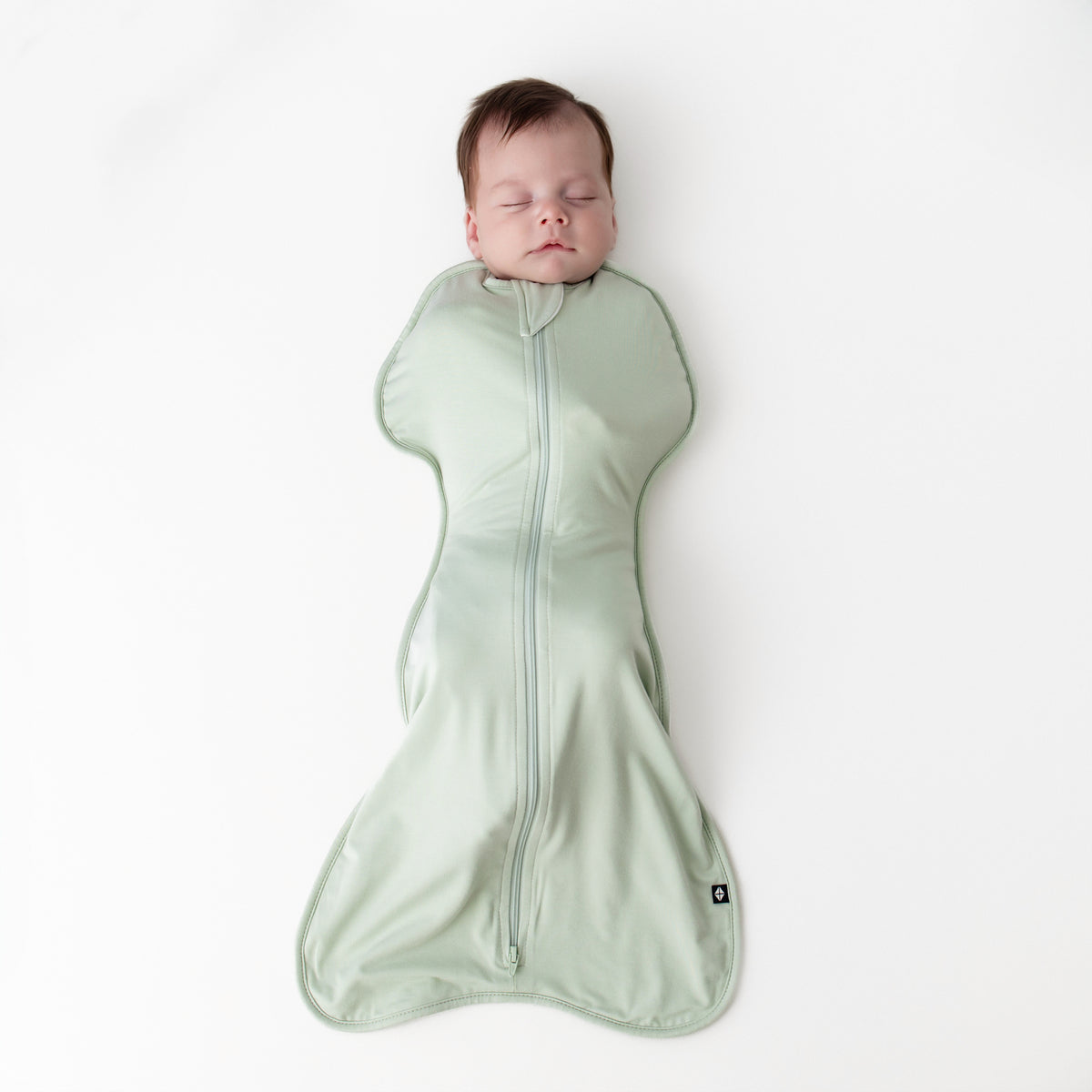 Sleeping newborn wearing the Swaddle Bag in Basil 0.5 laying on a white surface