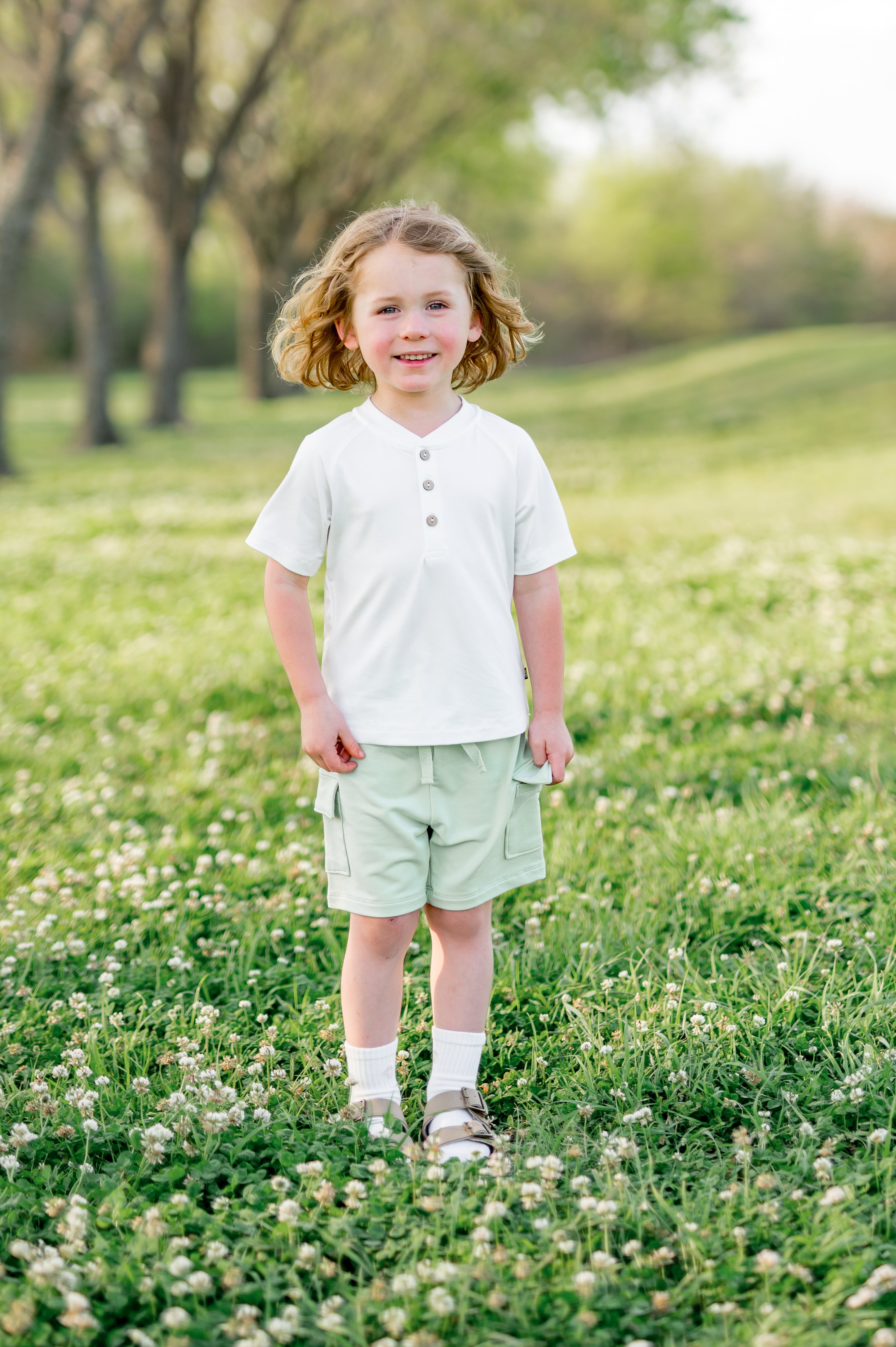 Young child standing in a grass field wearing the Cargo Short in Basil paired with a white short sleeve henley