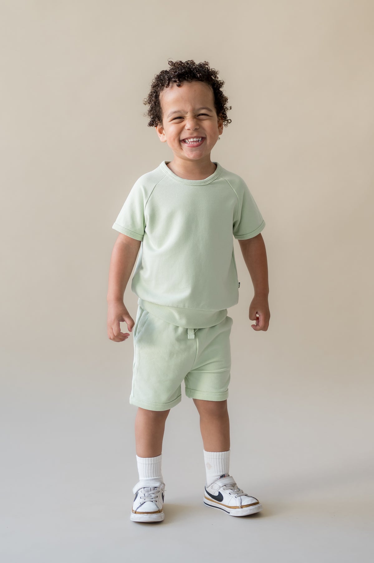 Smiling boy wearing the Short Sleeve Jogger Set in Basil in front of a light beige background
