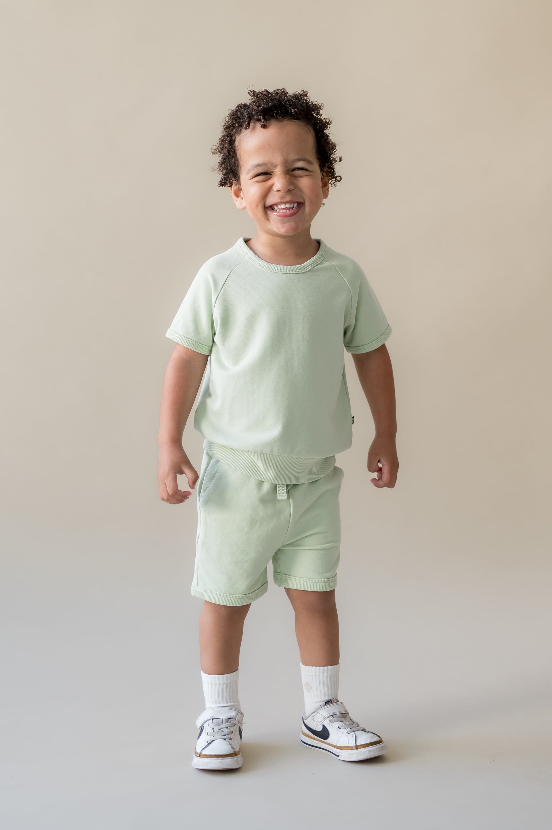 Smiling boy wearing the Short Sleeve Jogger Set in Basil in front of a light beige background