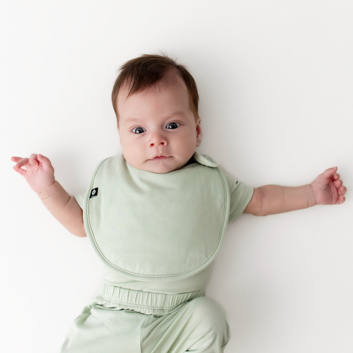 Newborn laying on a cream surface wearing the Cushy Bib in Basil with the matching short sleeve bodysuit and pants