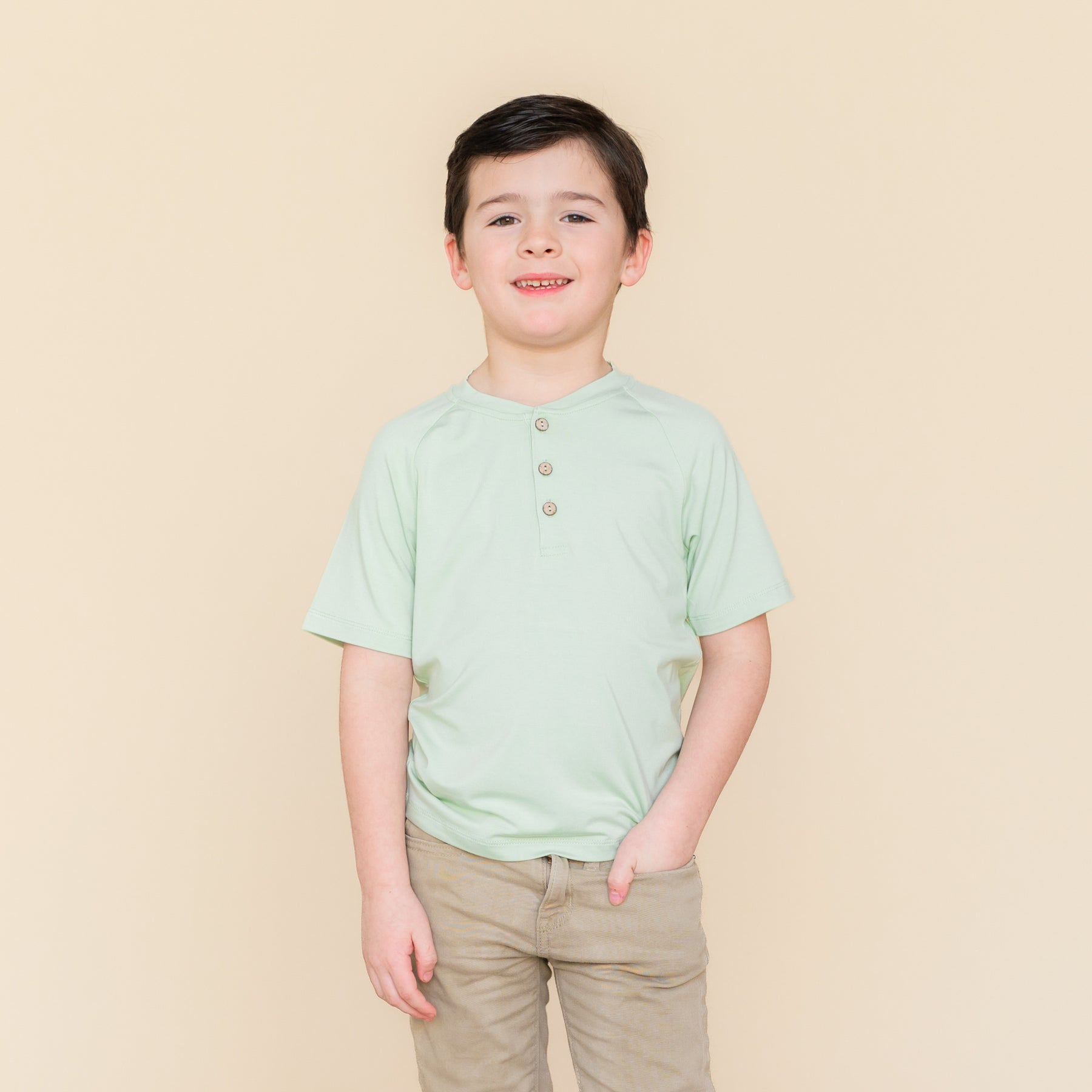 Young boy modeling the Short Sleeve Toddler Henley Tee in Basil with tan colored pants