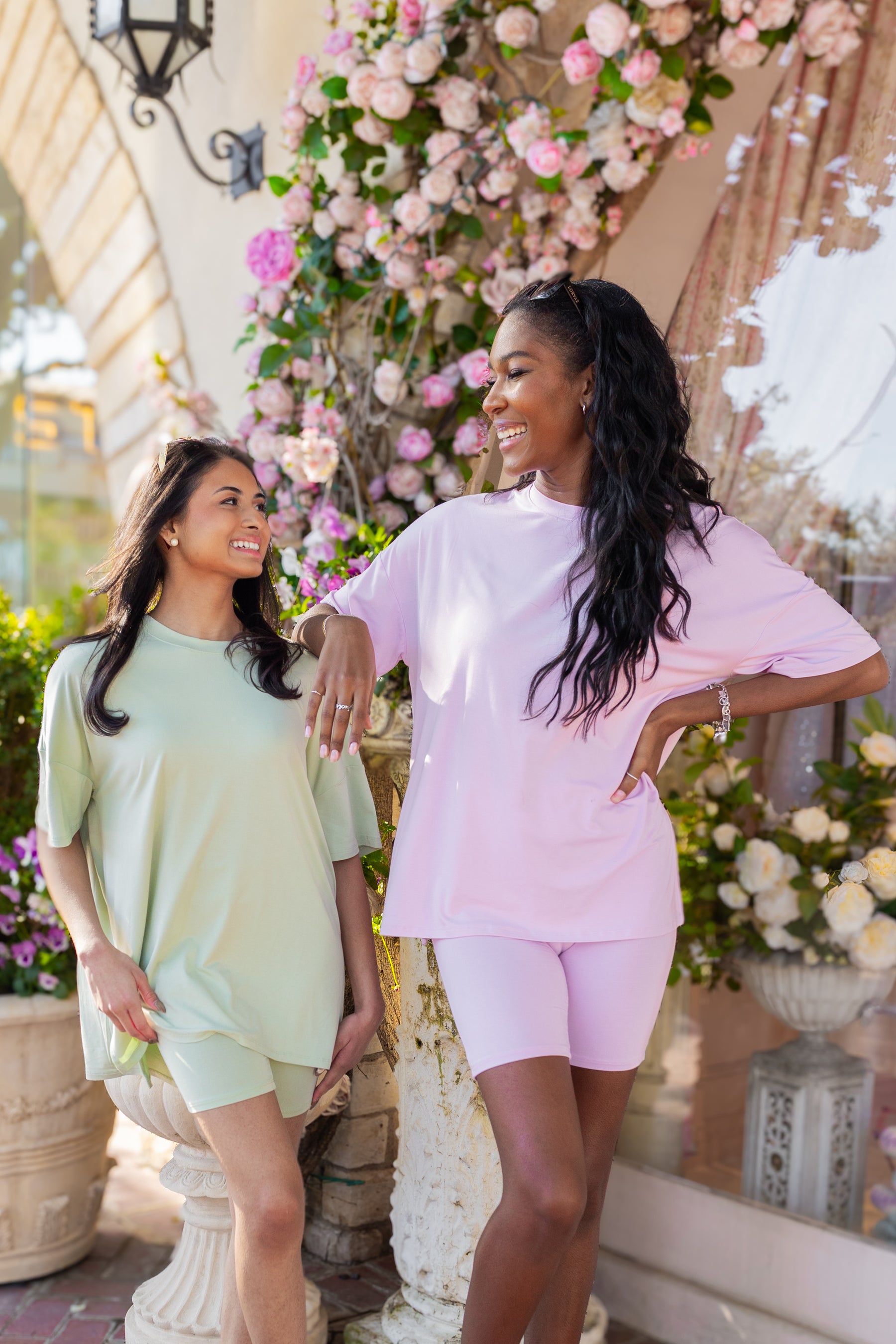Two women standing outdoors with floral decorations in the background wearing biker short sets