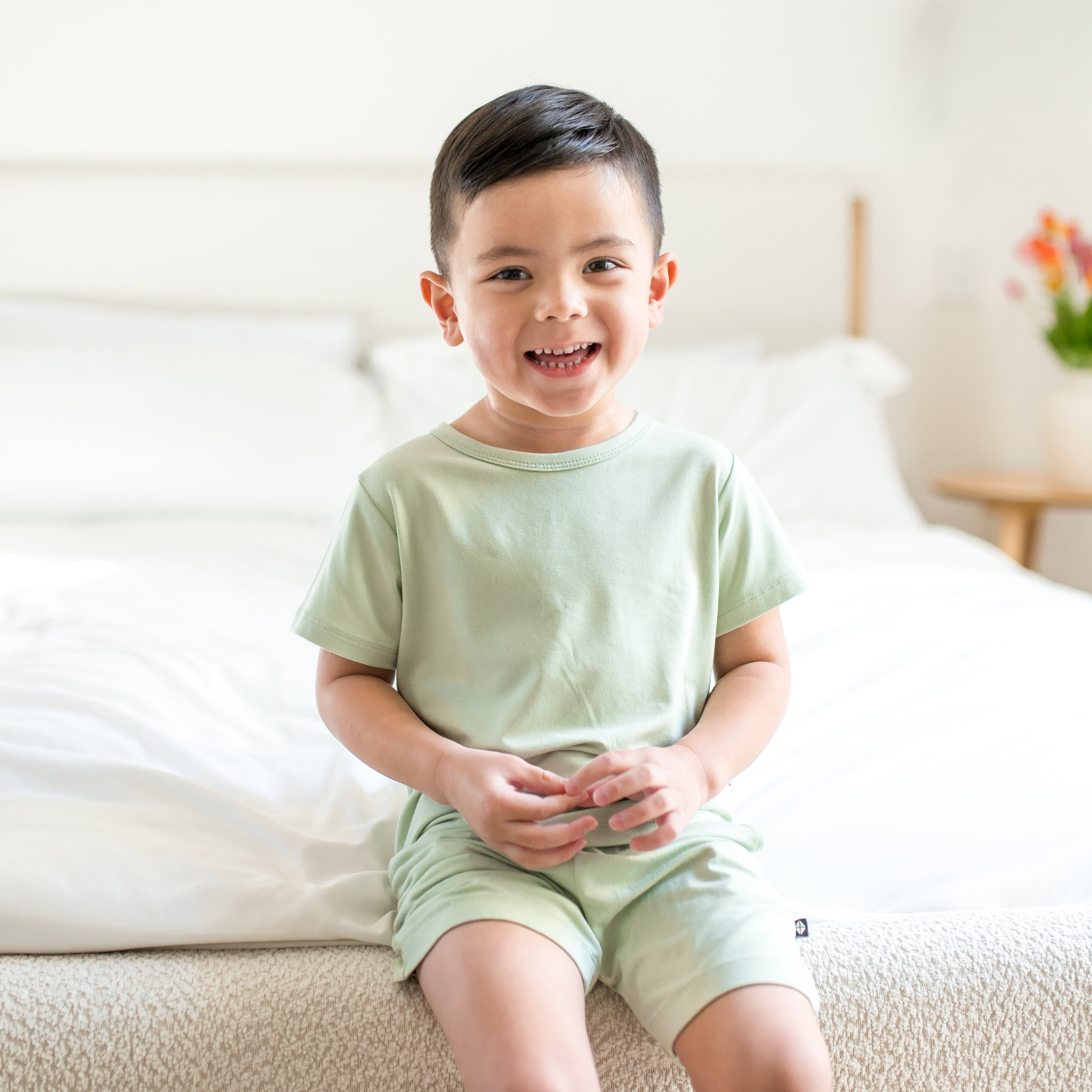 Young boy sitting on the end of the bed wearing the Toddler Basic Tee in Basil and matching shorts