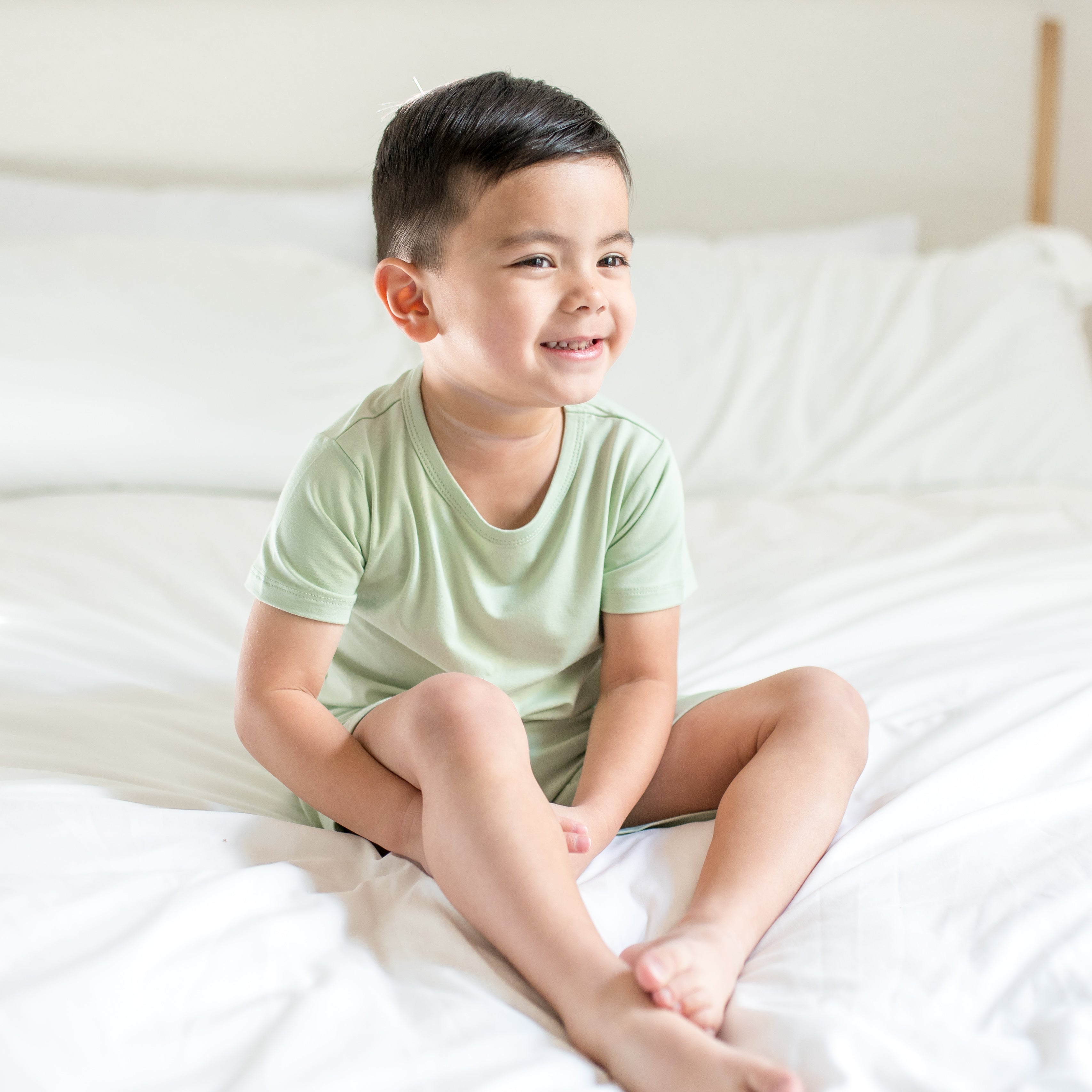 Young boy sitting on a bed wearing the Toddler Basic Tee in Basil