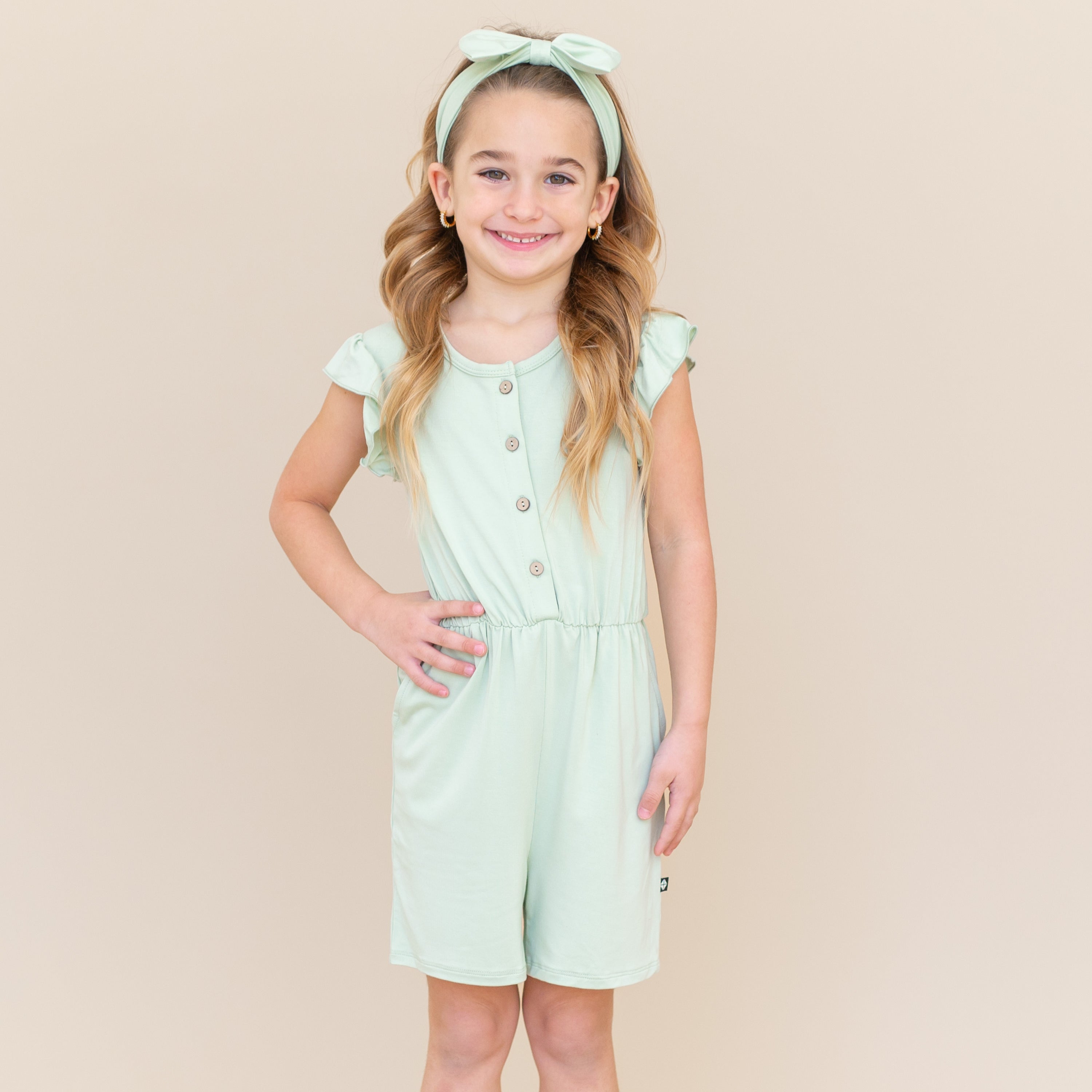 Smiling girl standing with one hand on her hip wearing the Flutter Sleeve Romper in Basil and matching bow
