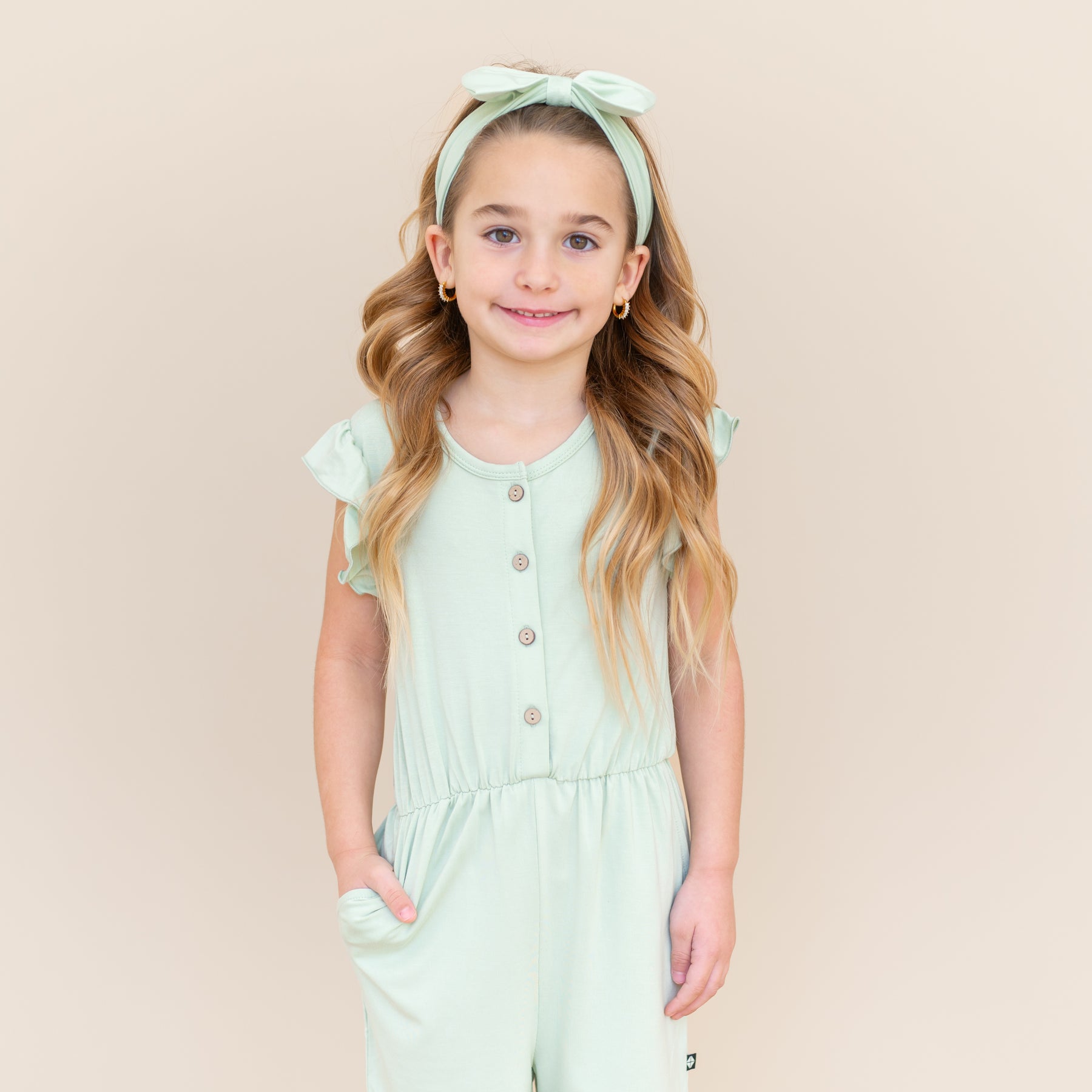 Close up of smiling girl wearing the Flutter Sleeve Romper in Basil with one hand in the romper pocket