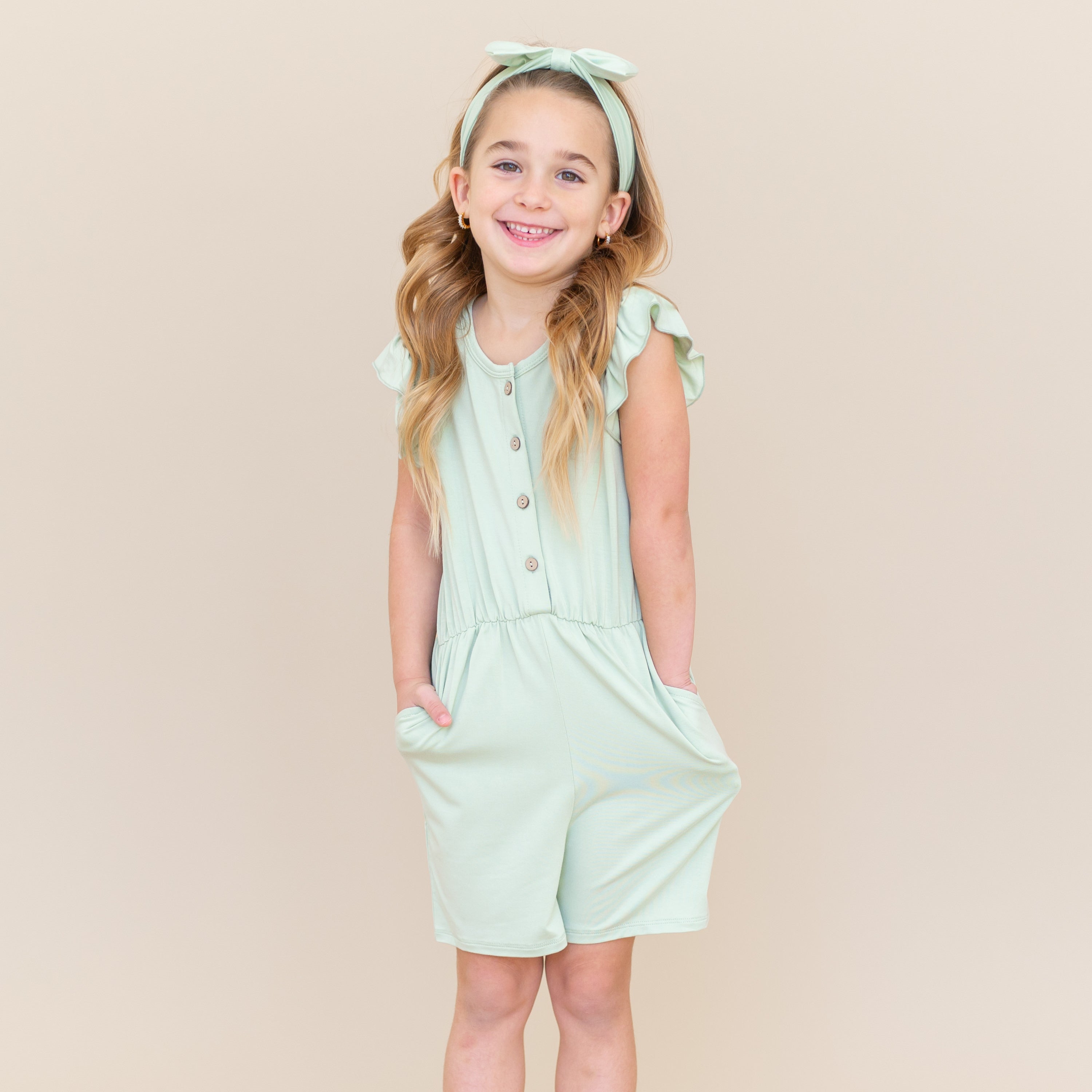 Smiling girl modeling the Flutter Sleeve Romper in Basil with her hands in the pockets