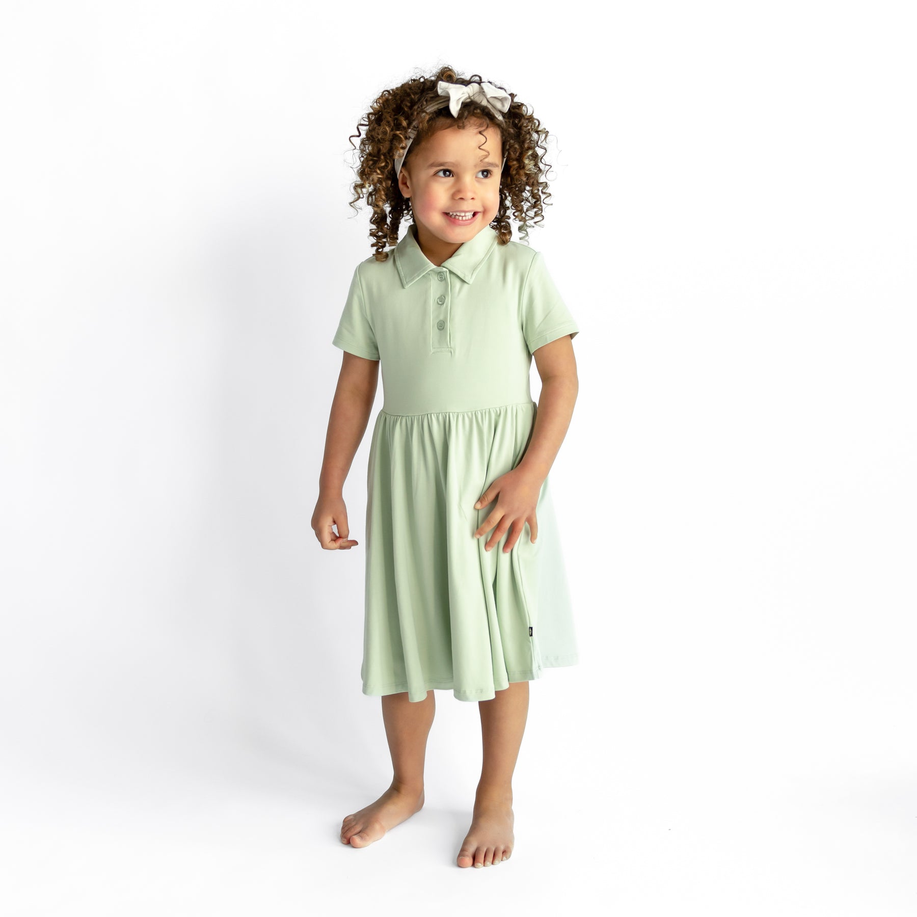 Young girl smiling wearing the Polo Dress in Basil standing in front of a white background