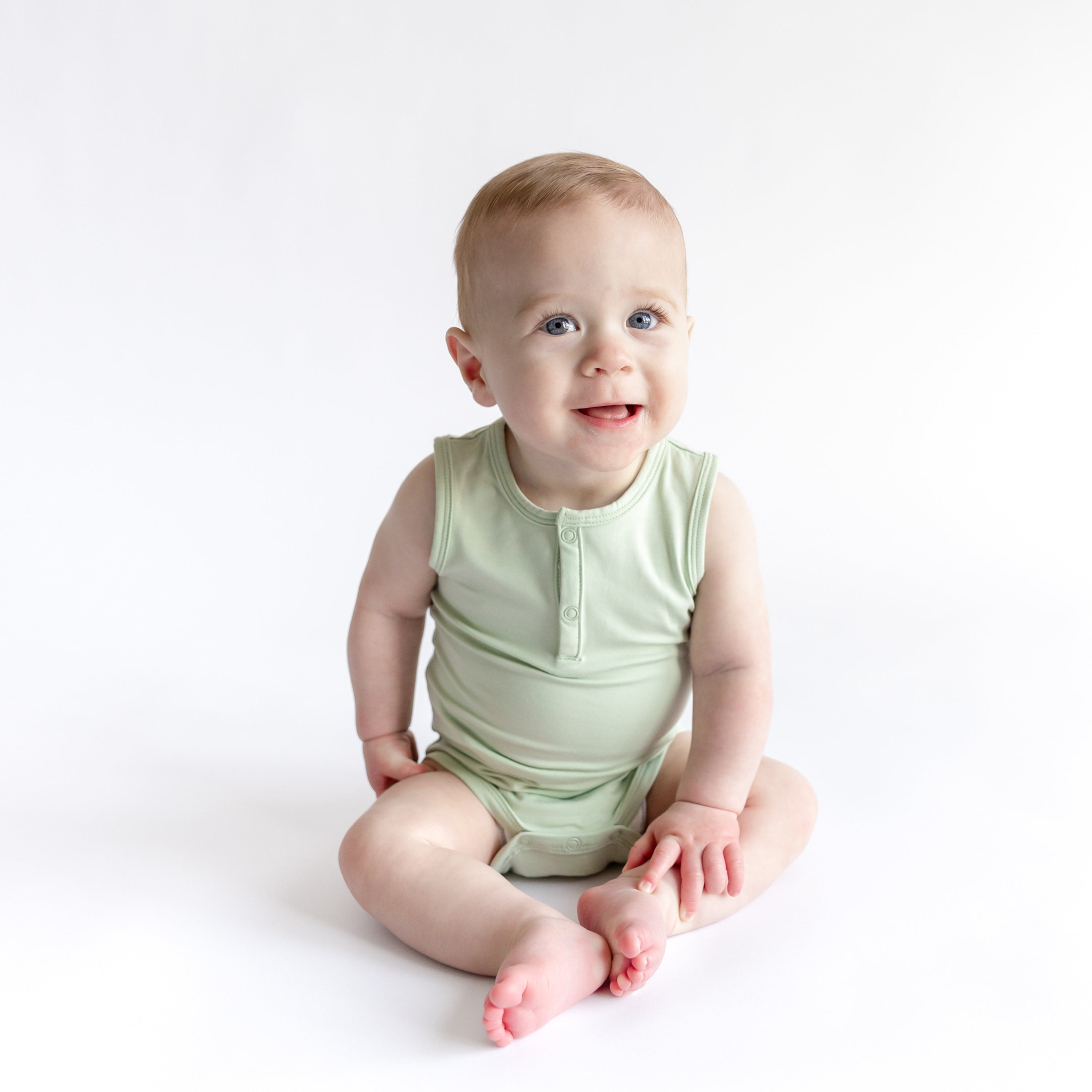 Smiling toddler sitting on the floor wearing the Sleeveless Bodysuit in Basil