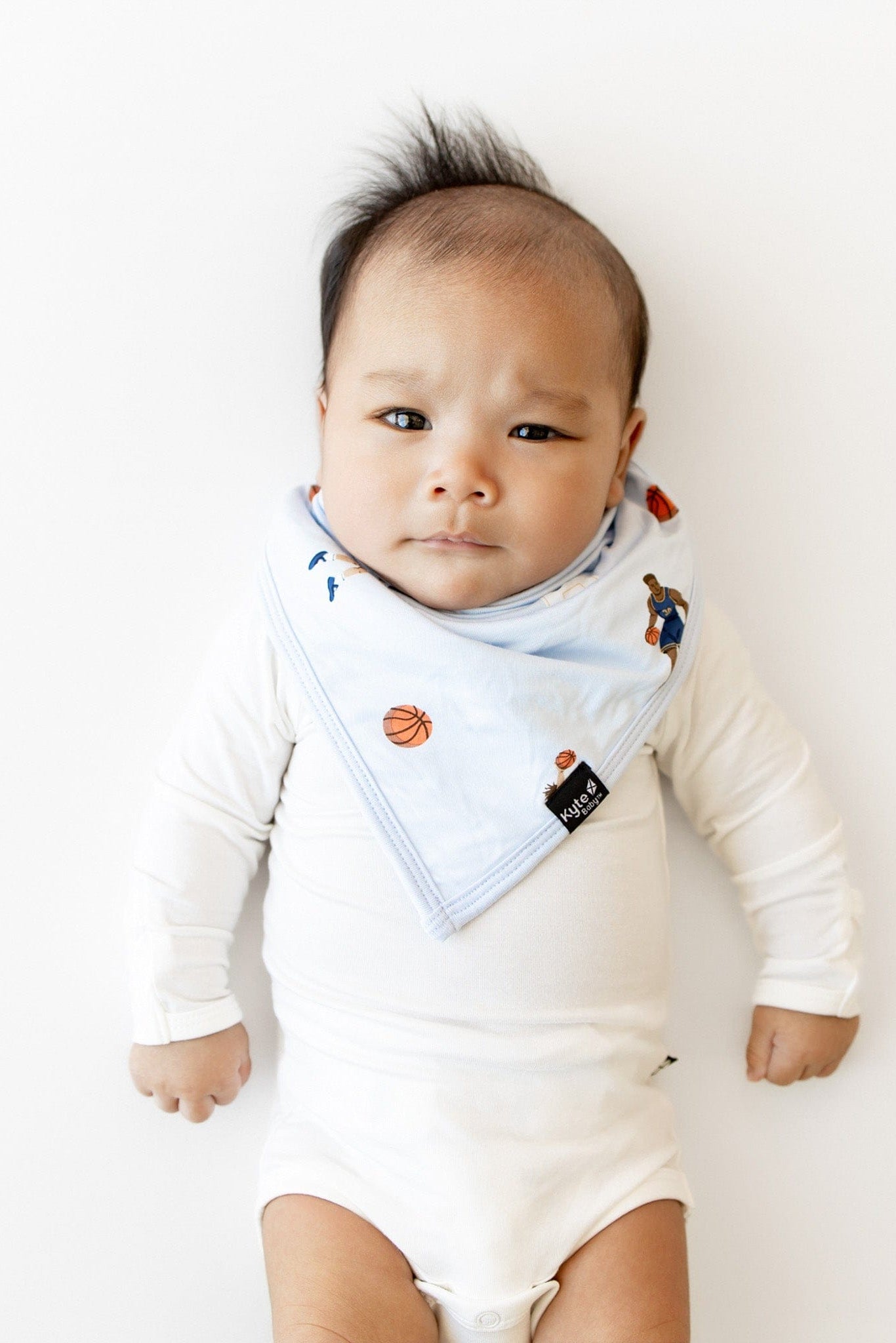 Infant laying on a white surface wearing the Bib in Basketball overtop a white long sleeve bodysuit