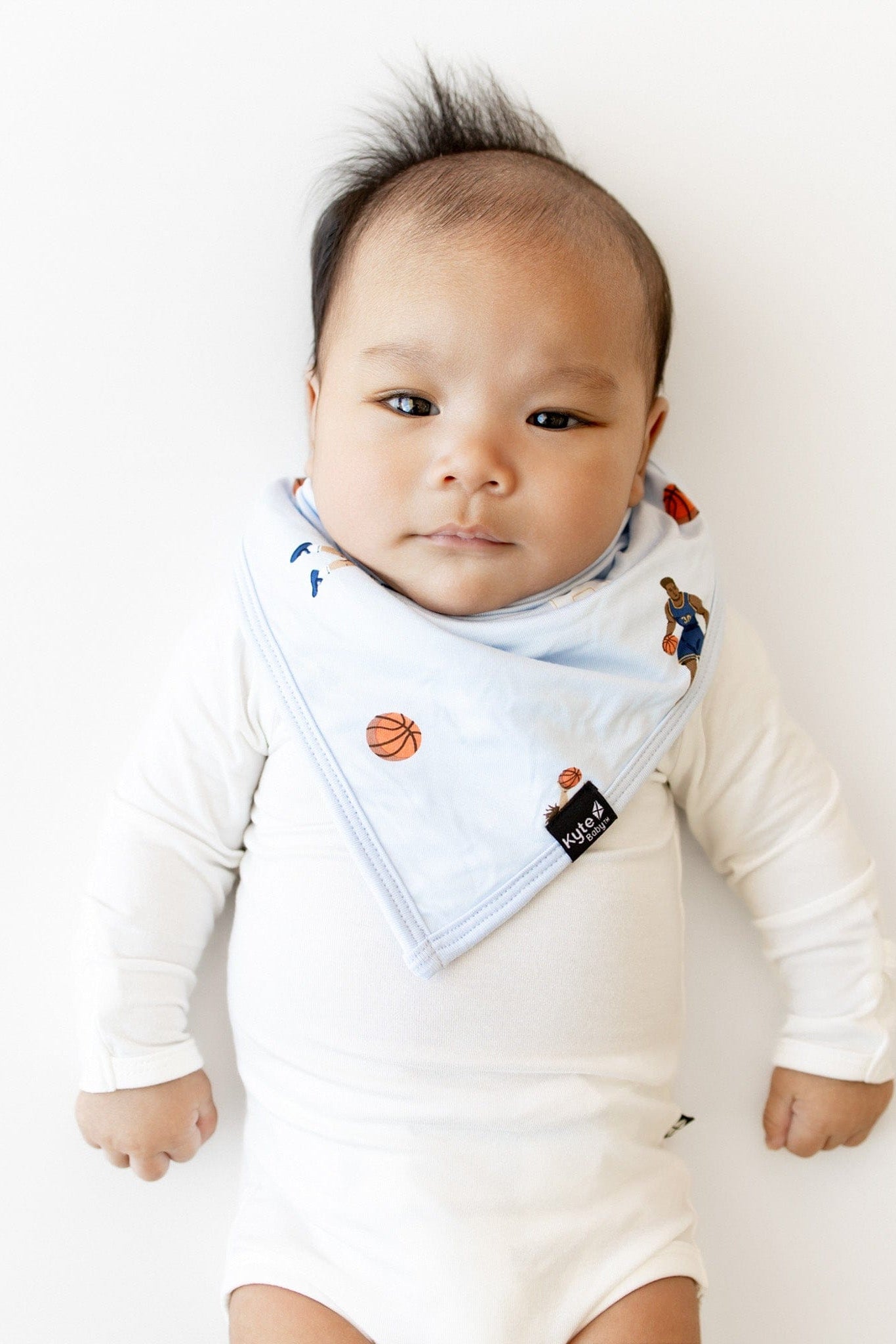 Infant laying in a white surface wearing the Bib in Basketball overtop a white long sleeve bodysuit