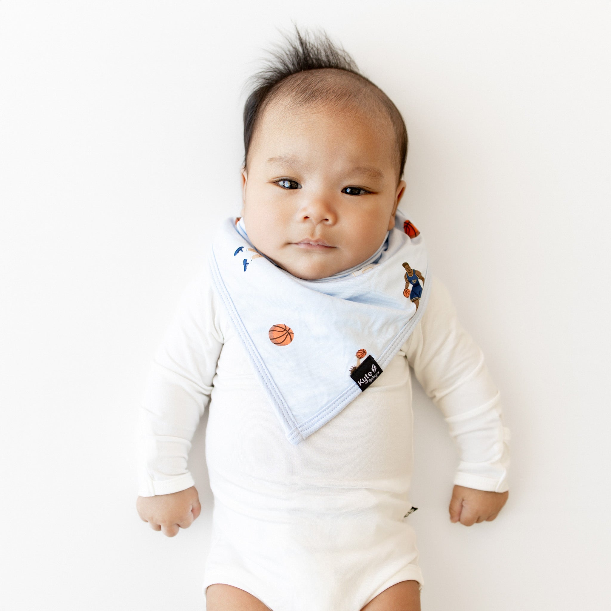 Infant laying in a white surface wearing the Bib in Basketball overtop a white long sleeve bodysuit