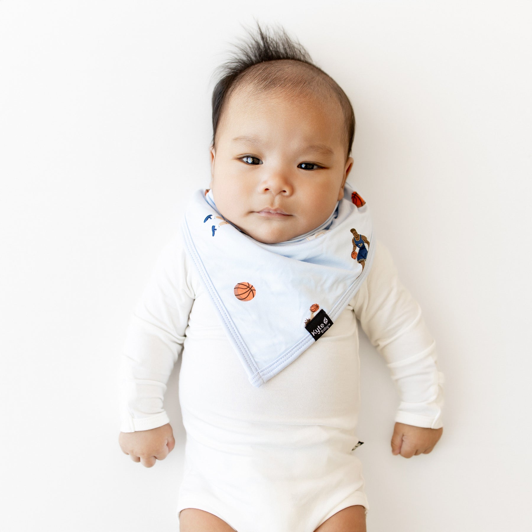 Infant laying in a white surface wearing the Bib in Basketball overtop a white long sleeve bodysuit