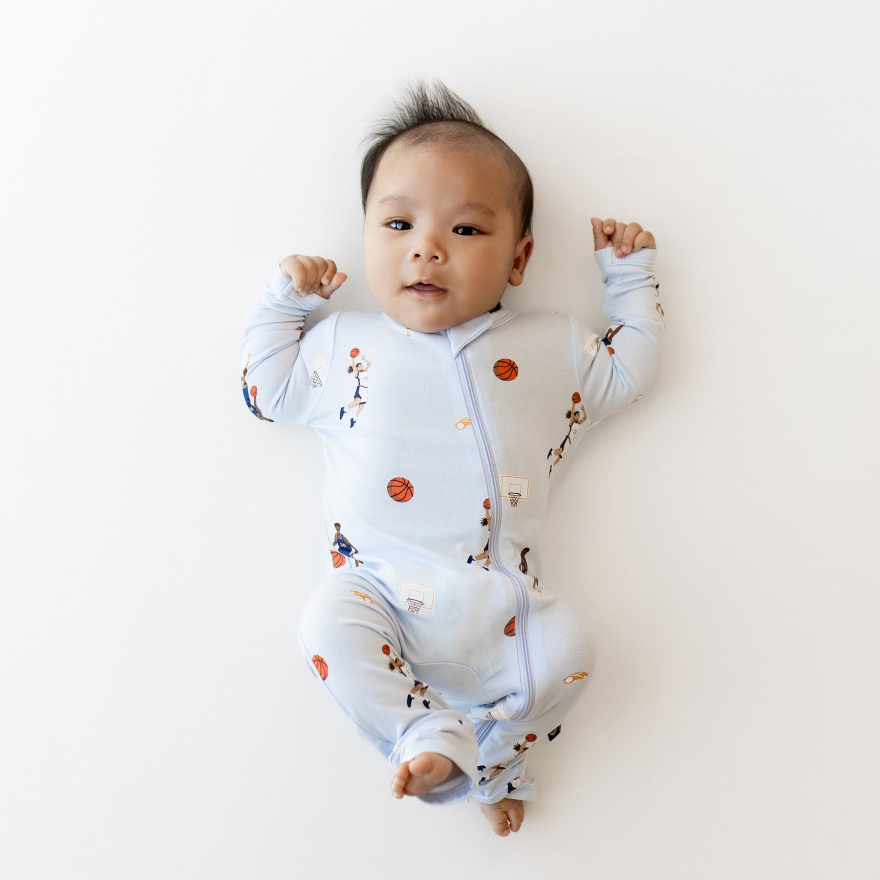 Infant laying on a light neutral surface wearing the Zippered Romper in Basketball