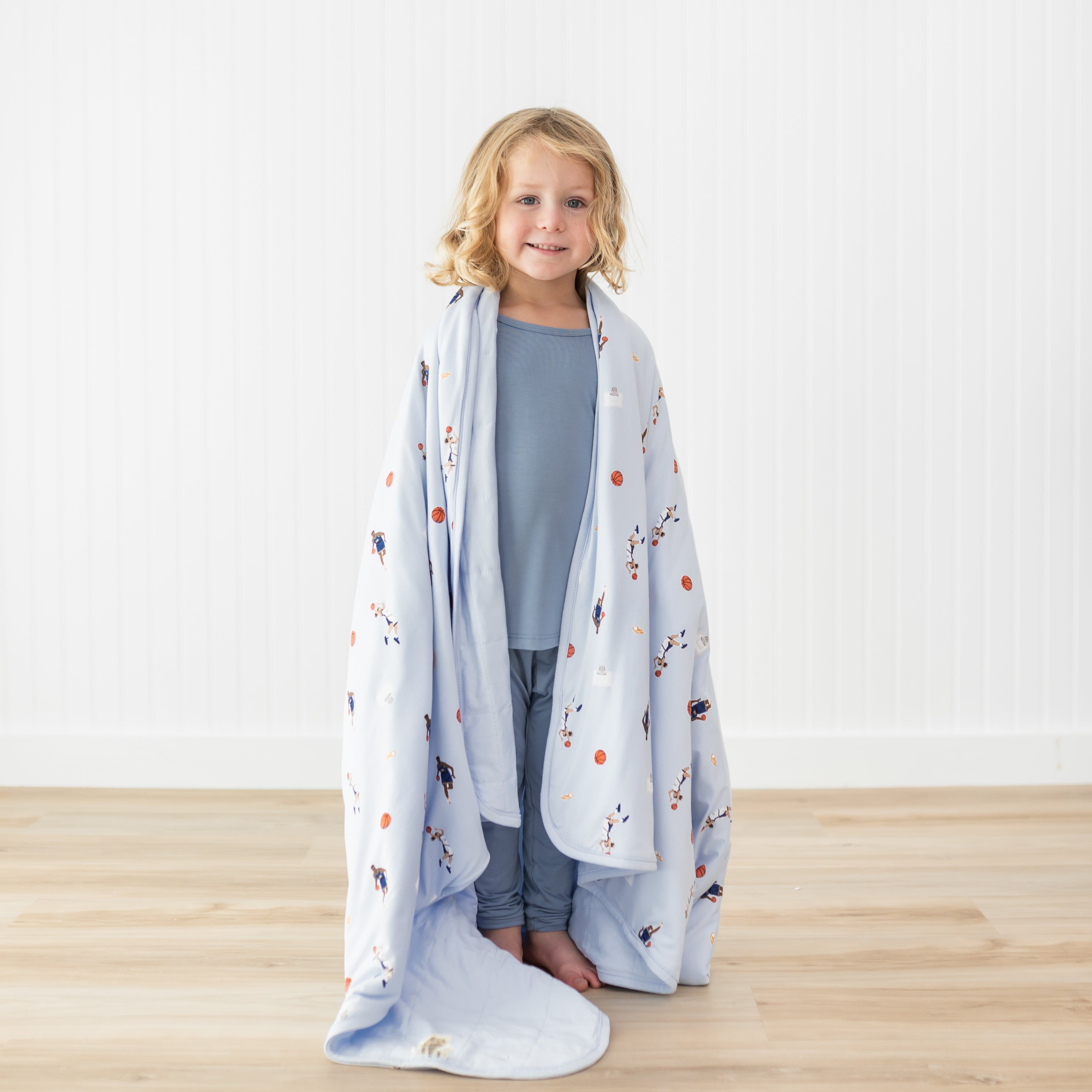 Young model standing wearing mute blue pajamas with the Toddler Blanket in Basketball 1.0 around their shoulders