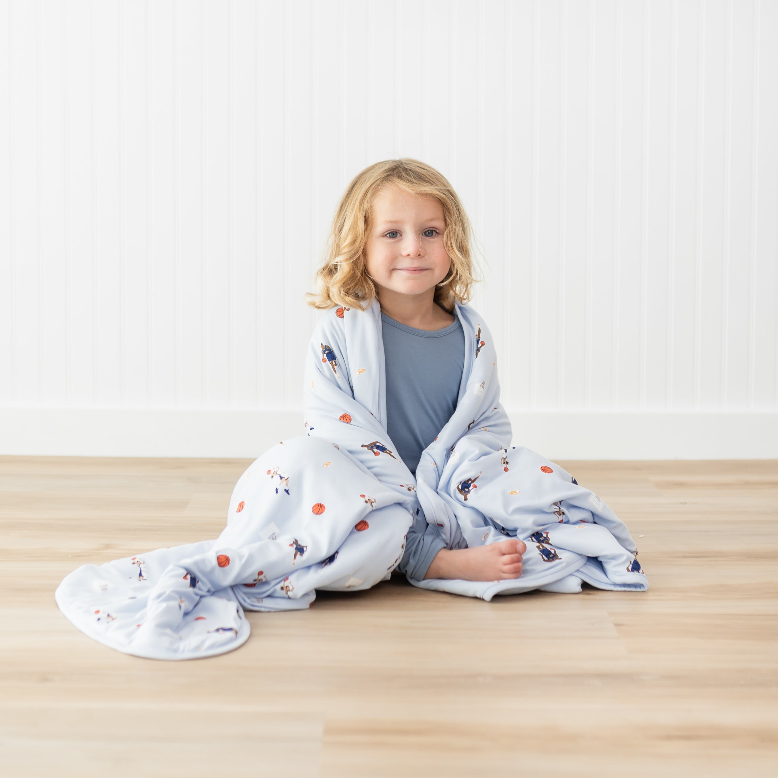 Young model sitting on the floor with the Toddler Blanket in Basketball 1.0 around their shoulders