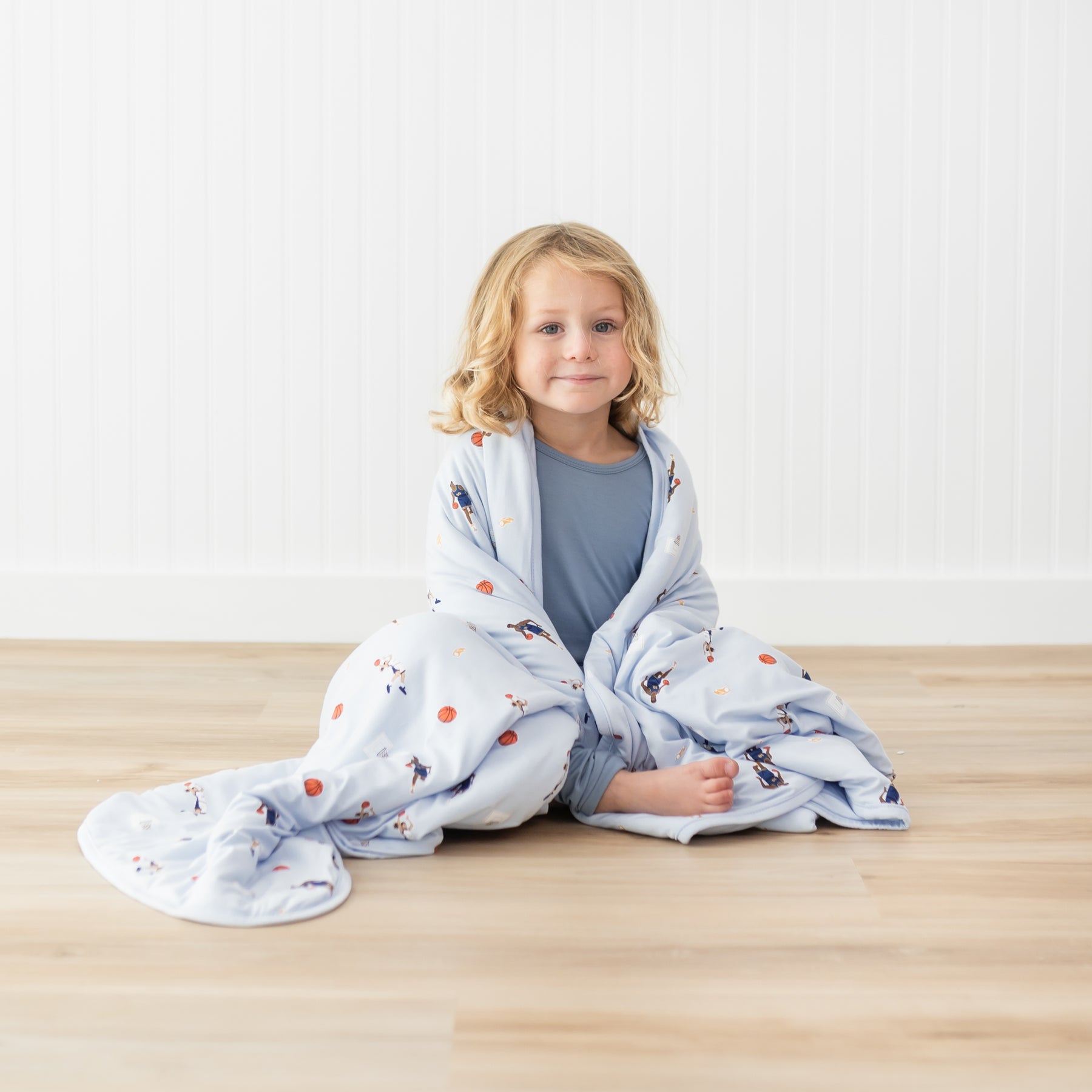 Young model sitting on the floor with the Toddler Blanket in Basketball 1.0 around their shoulders