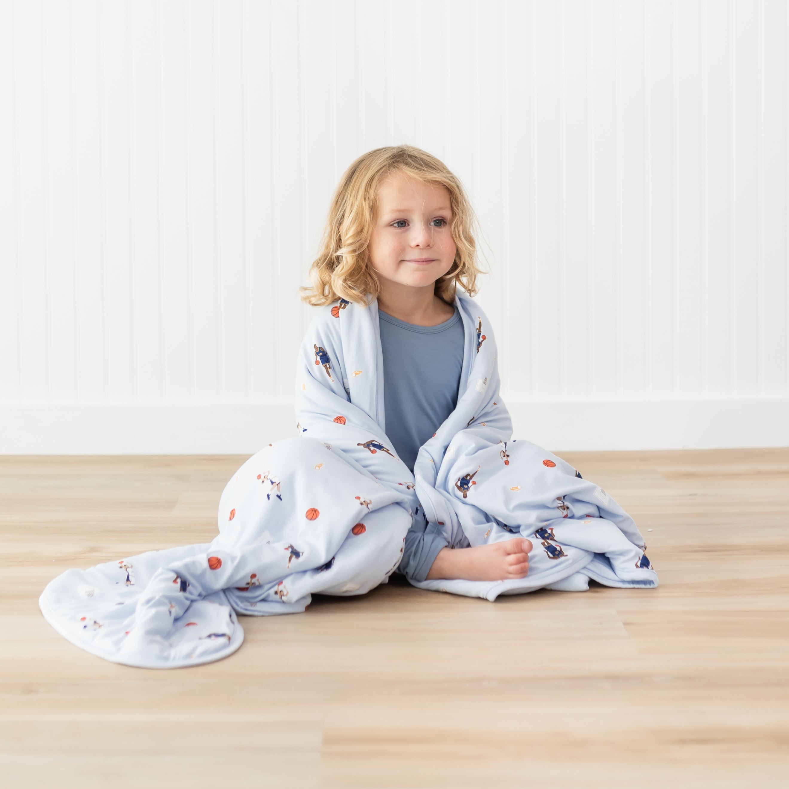 Young model sitting on the floor wearing muted blue pajamas with the Toddler Blanket in Basketball 1.0 around their shoulders