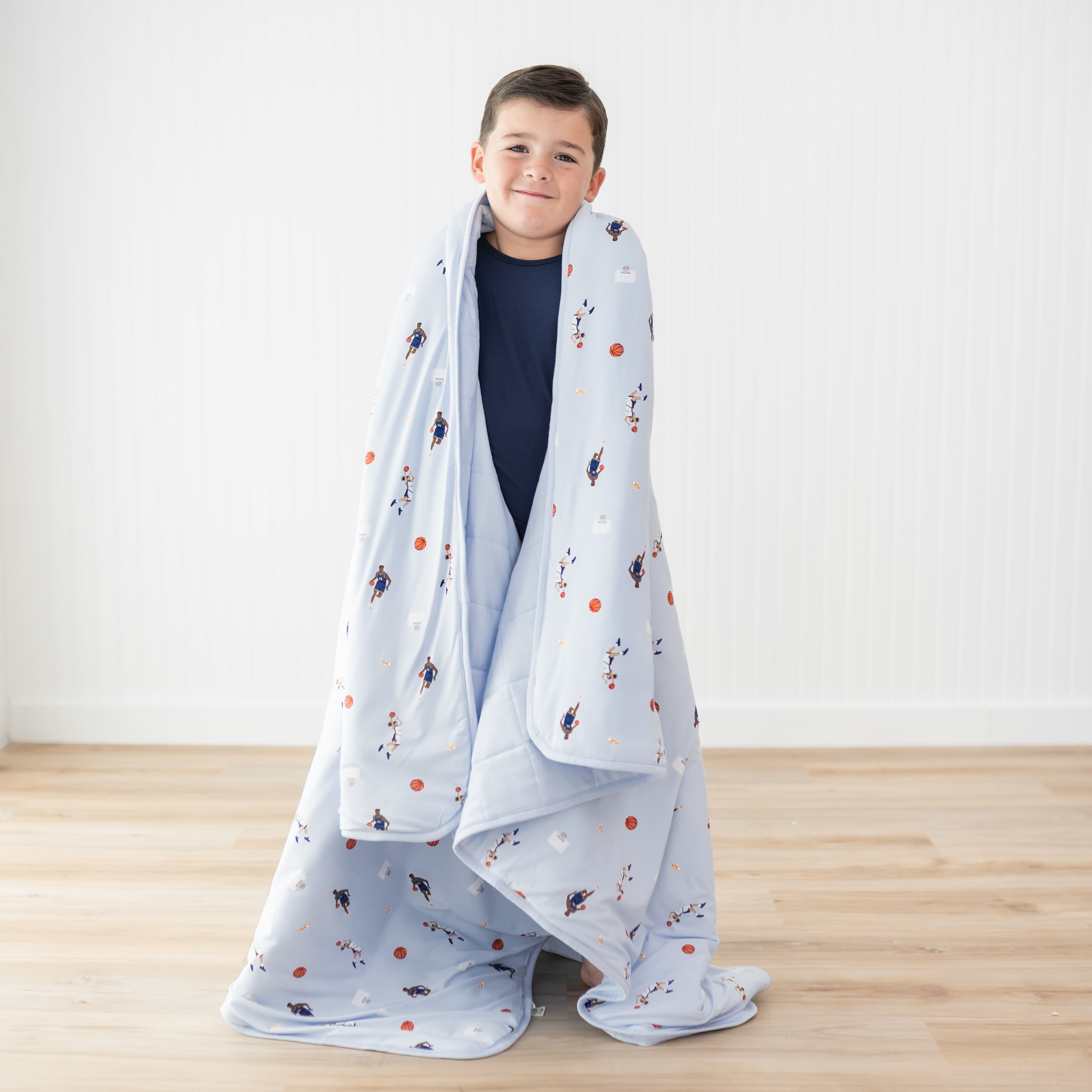 Young boy wearing dark blue long sleeve pajamas with the Youth Blanket in Basketball 2.5 overtop of his shoulders