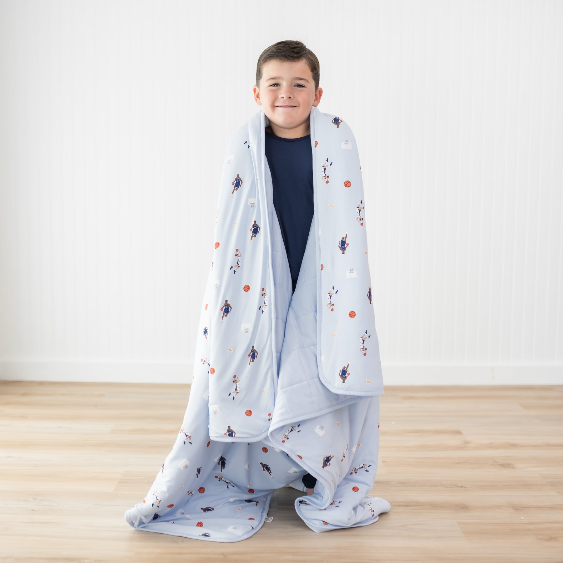 Young boy wearing dark blue pajamas standing with the Youth Blanket in Basketball 2.5 around his shoulders