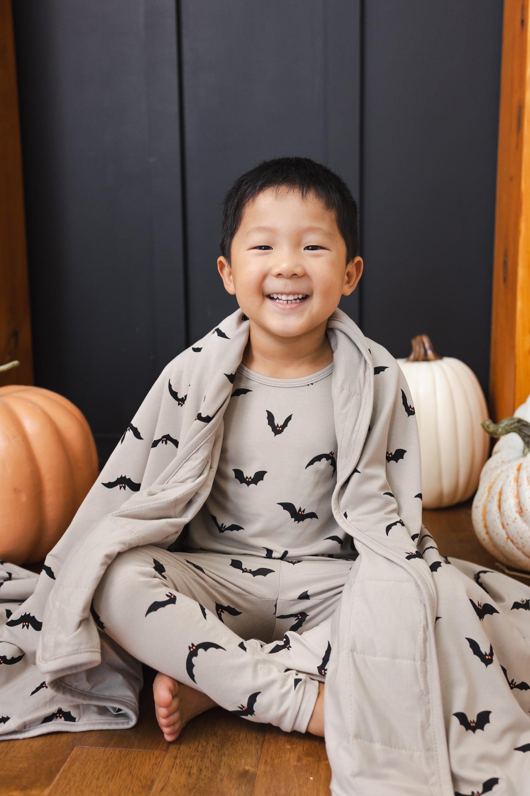 Boy wrapped in a Toddler Blanket in Bats 1.0 print sitting wearing a similar print long sleeve pajama set with pumpkin props all around him