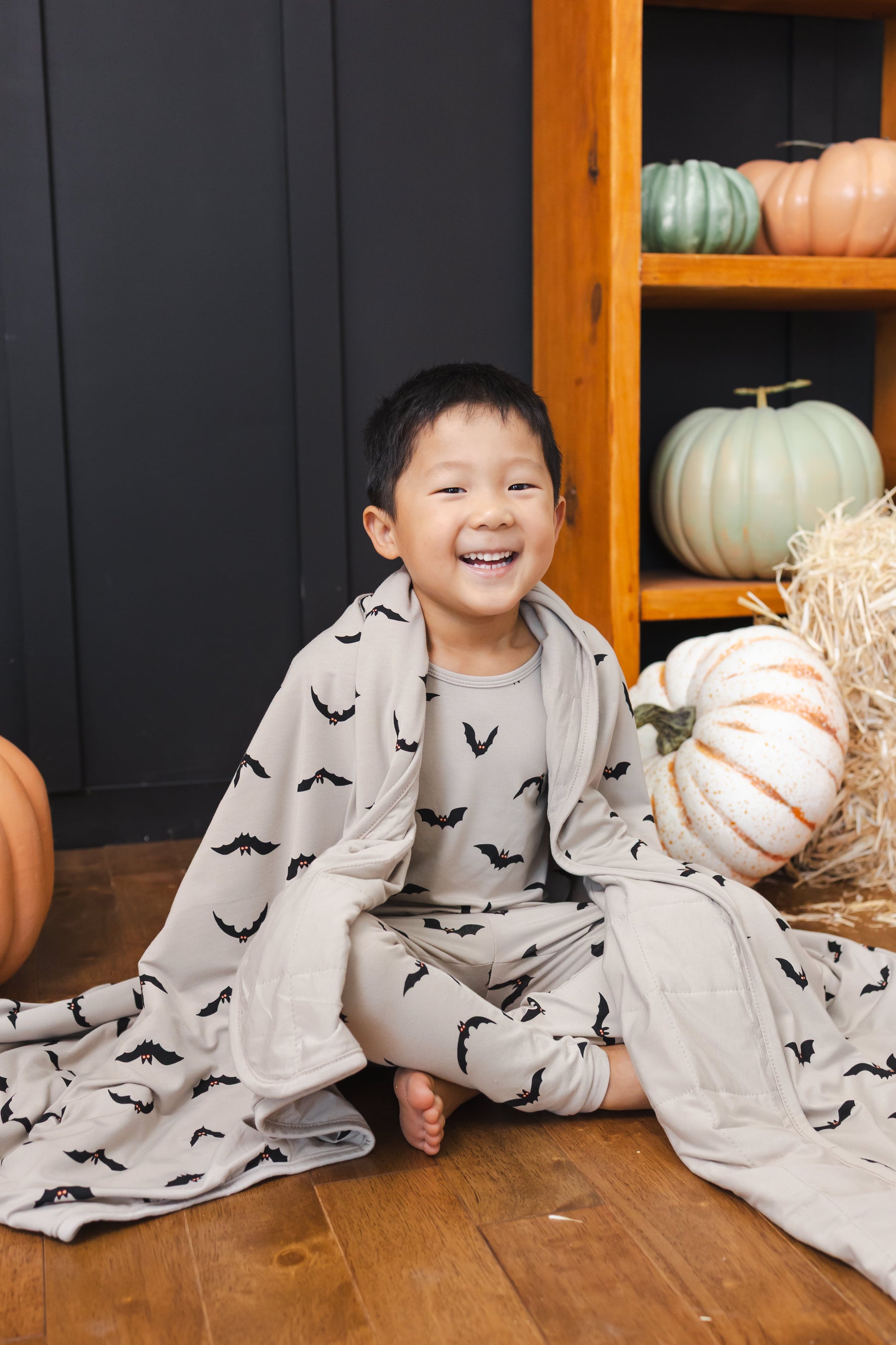 Boy smiling with a Toddler Blanket in Bats 1.0 print sitting wearing a similar print long sleeve pajama set with pumpkin all around him