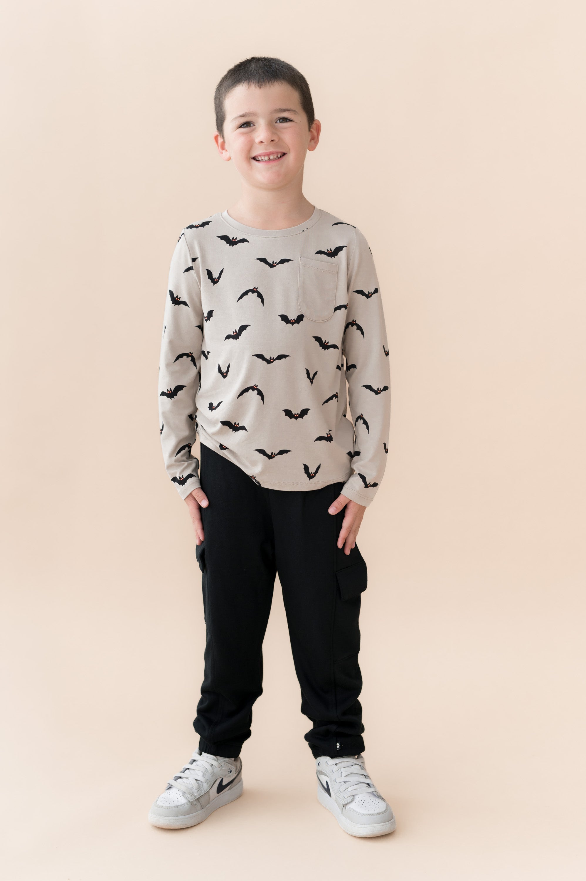 Boy posing in Long Sleeve Toddler Crew Neck Tee in Bats print over black pants and white sneakers on a beige background