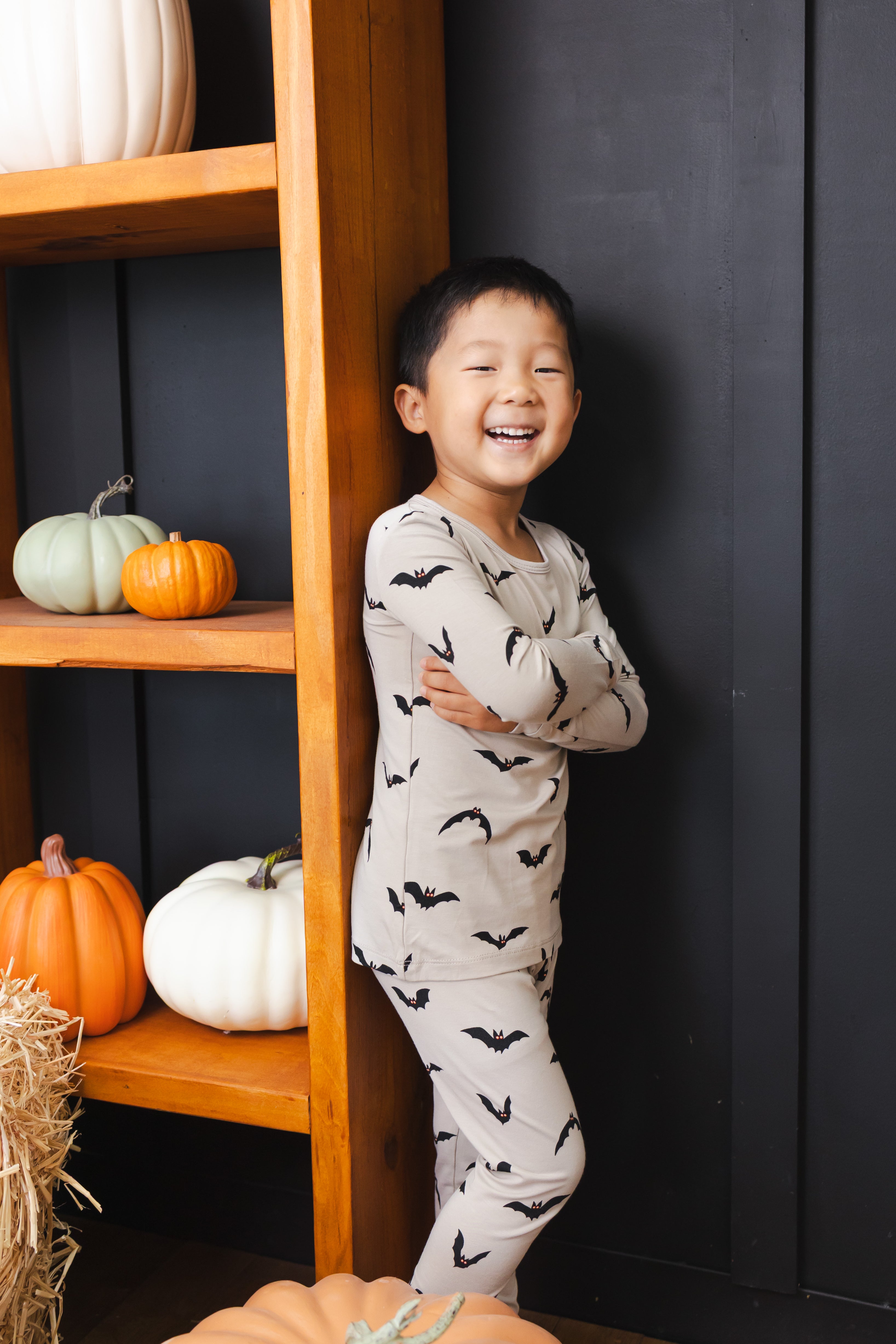 Boy smiling Long Sleeve Pajamas in Bats print standing against a shelf with pumpkin decorations