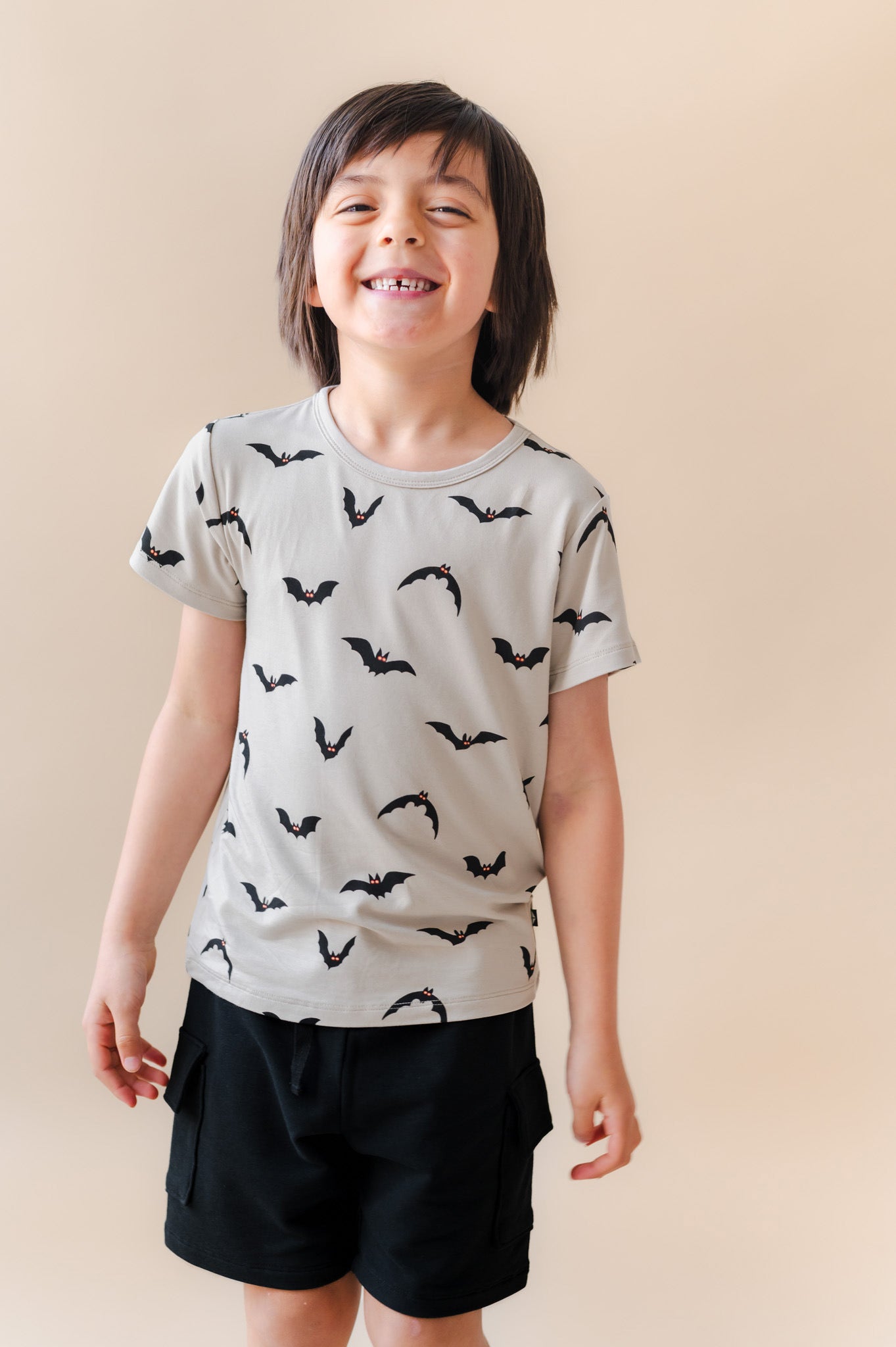 Boy smiling in Toddler Basic Tee in Bats print over black shorts on a beige background