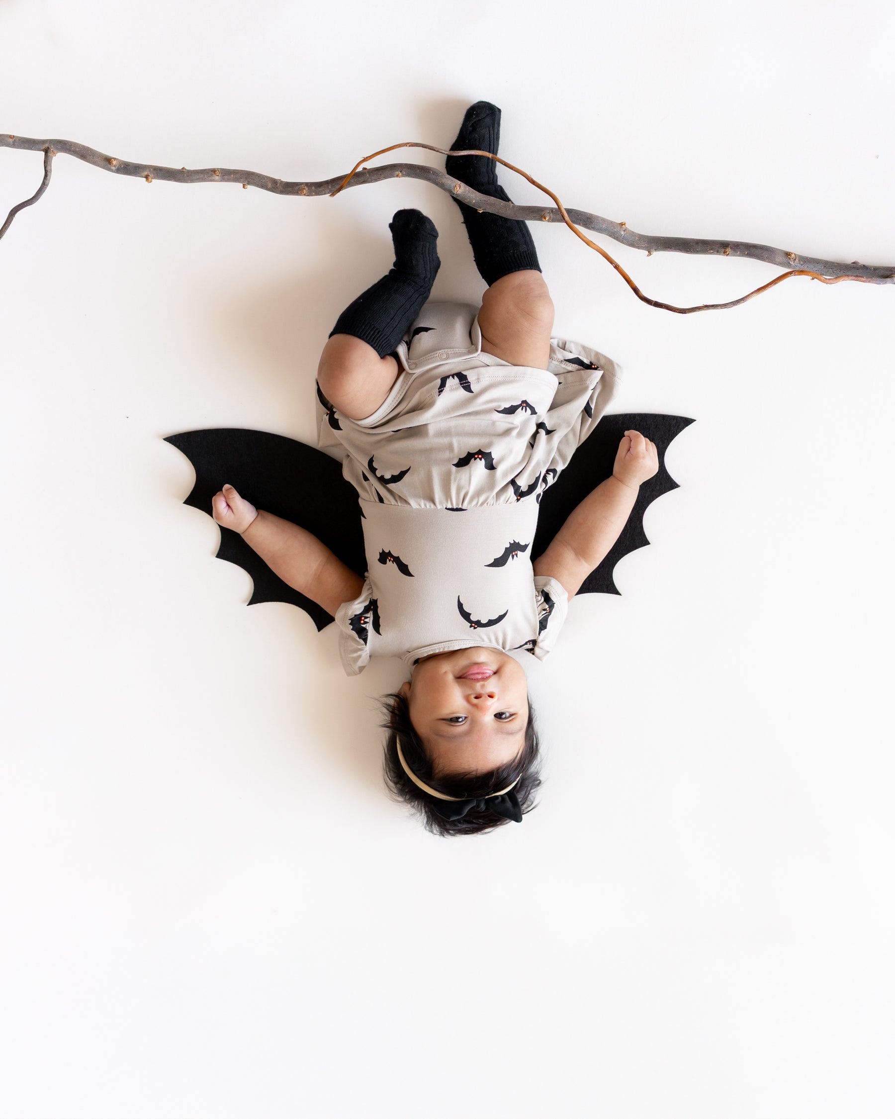 Baby posing as a bat hanging upside down from a branch wearing Twirl Bodysuit Dress in Bats print with bat wings and black socks on a white background