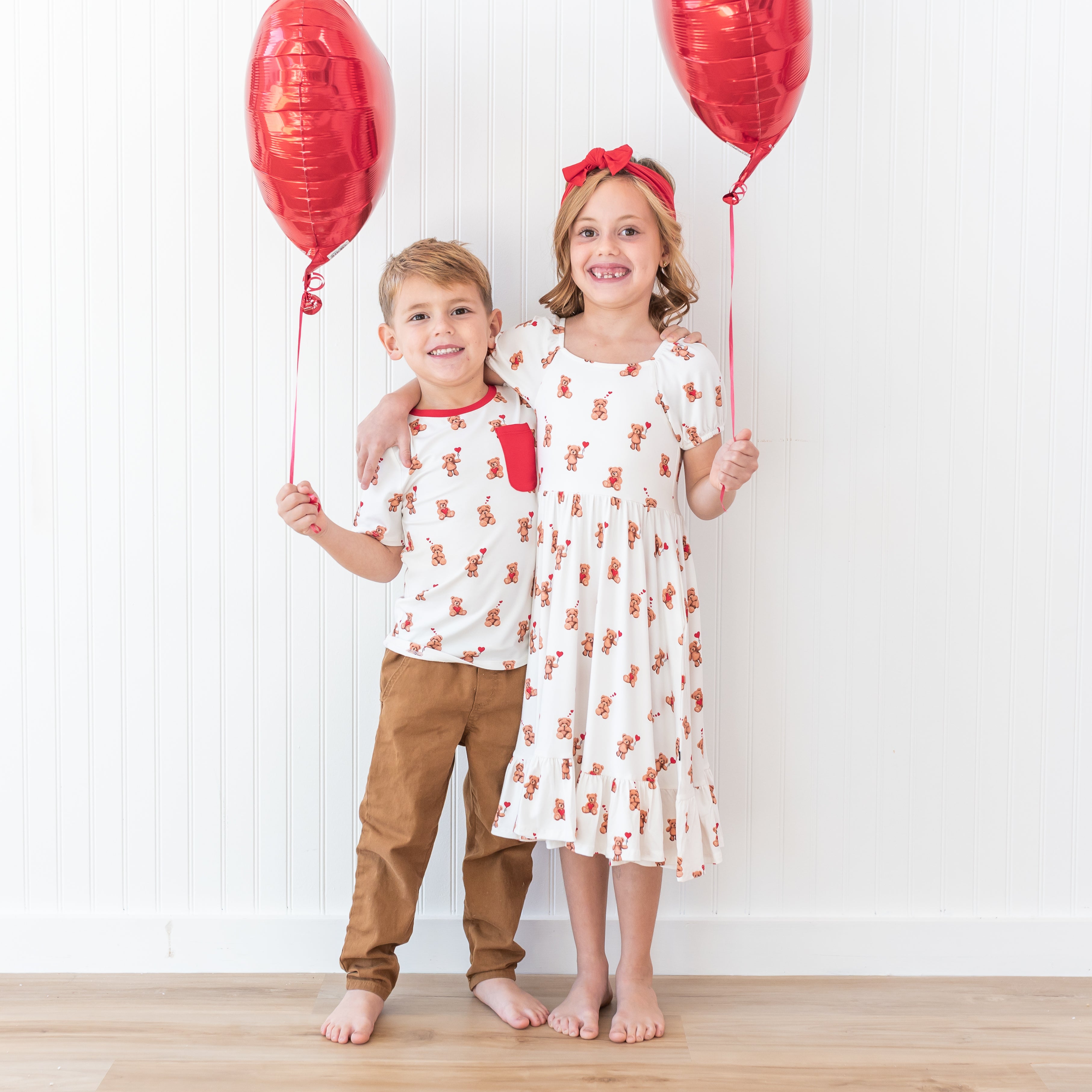 Brother and sister standing side by side with their arms around one another holding red heart balloons wearing the Toddler Crew Neck Tee in Bear Hearts and puff sleeve twirl dress in bear hearts