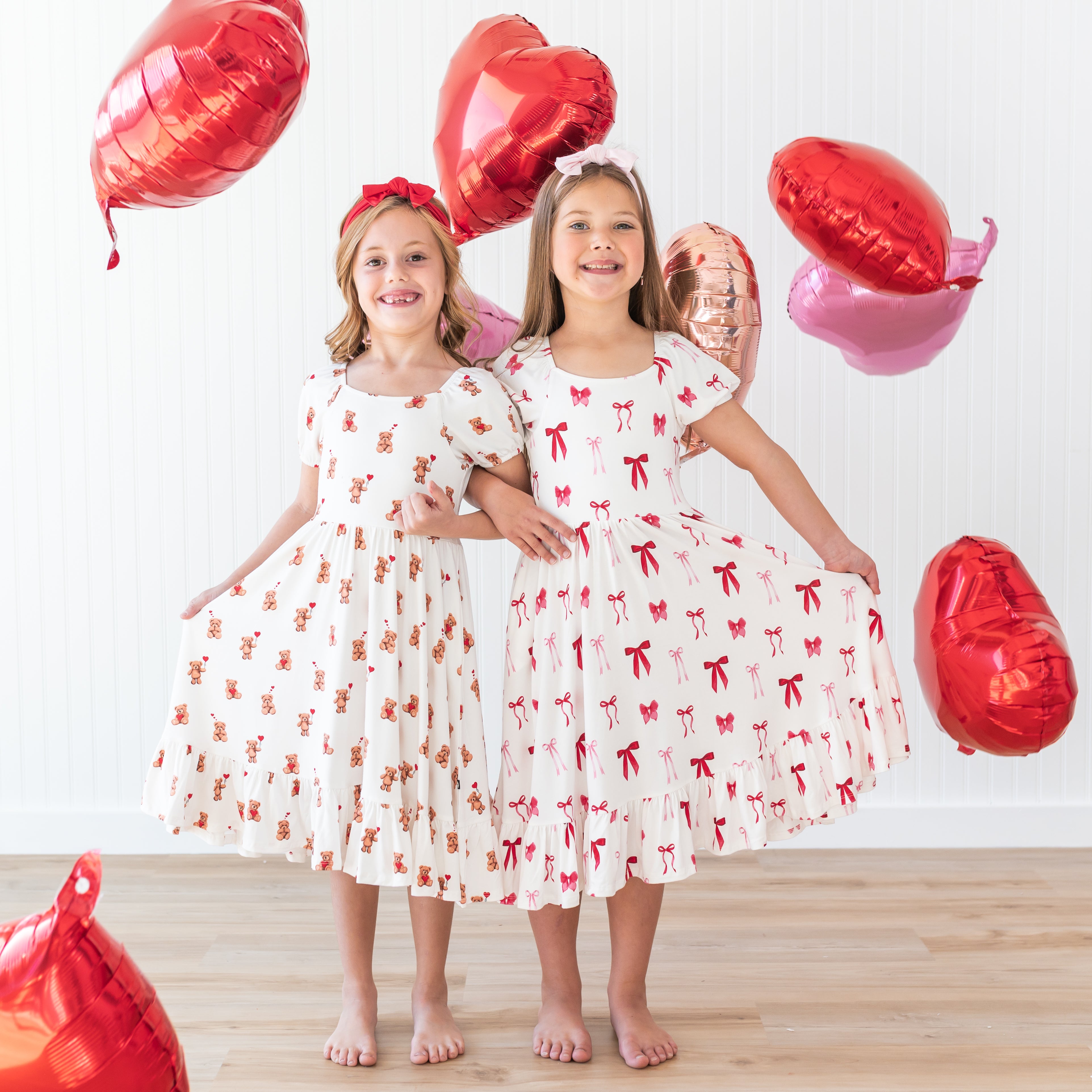 Two girls standing side by side with arms lined wearing the Puff Sleeve Twirl Dresses in Bear Hearts and Small Love Bow surrounded by red and pink heart balloons