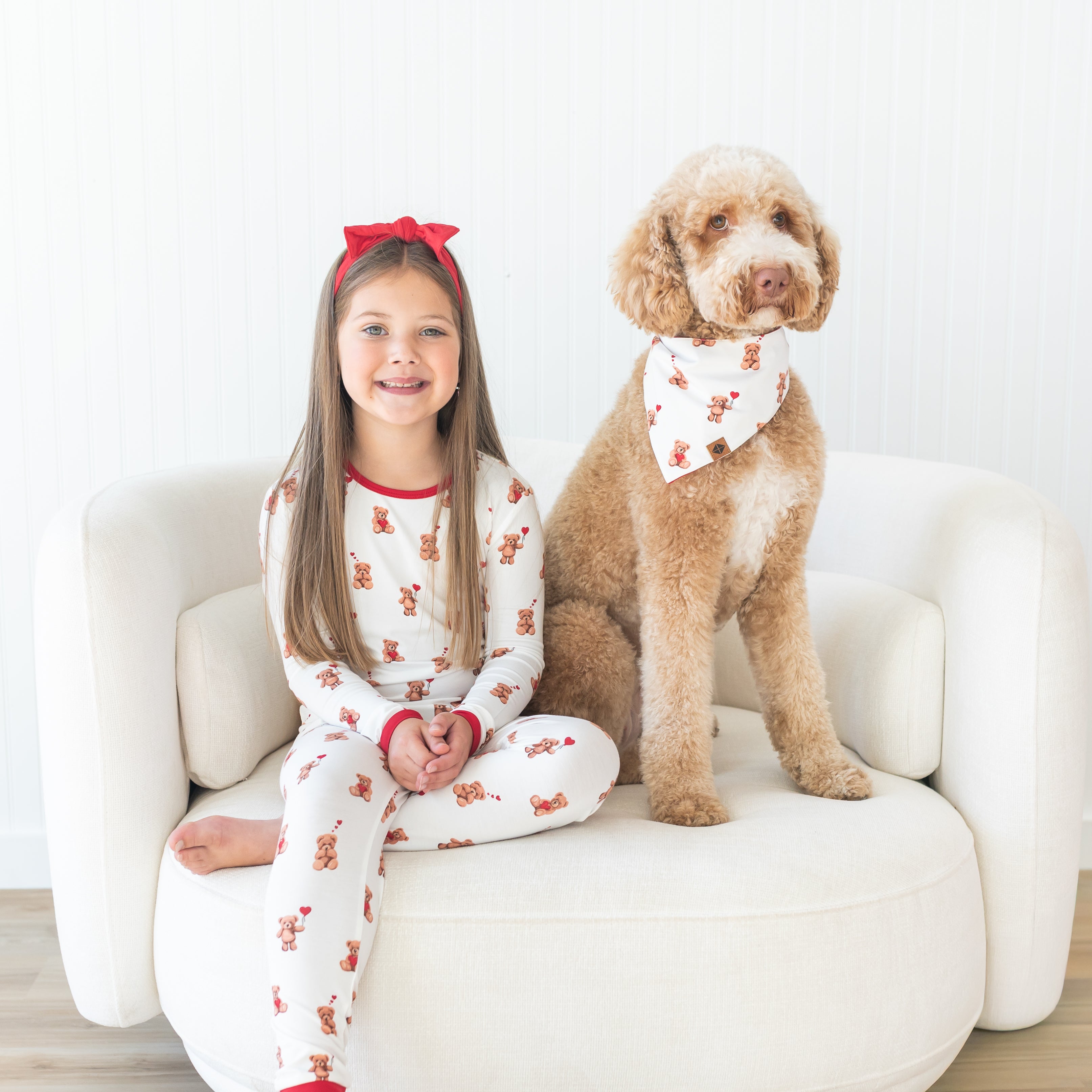Young girl wearing the Long Sleeve Pajamas in Bear Hearts sitting on a round cream sofa beside her pet dog who is wearing a matching dog bandana in bear hearts