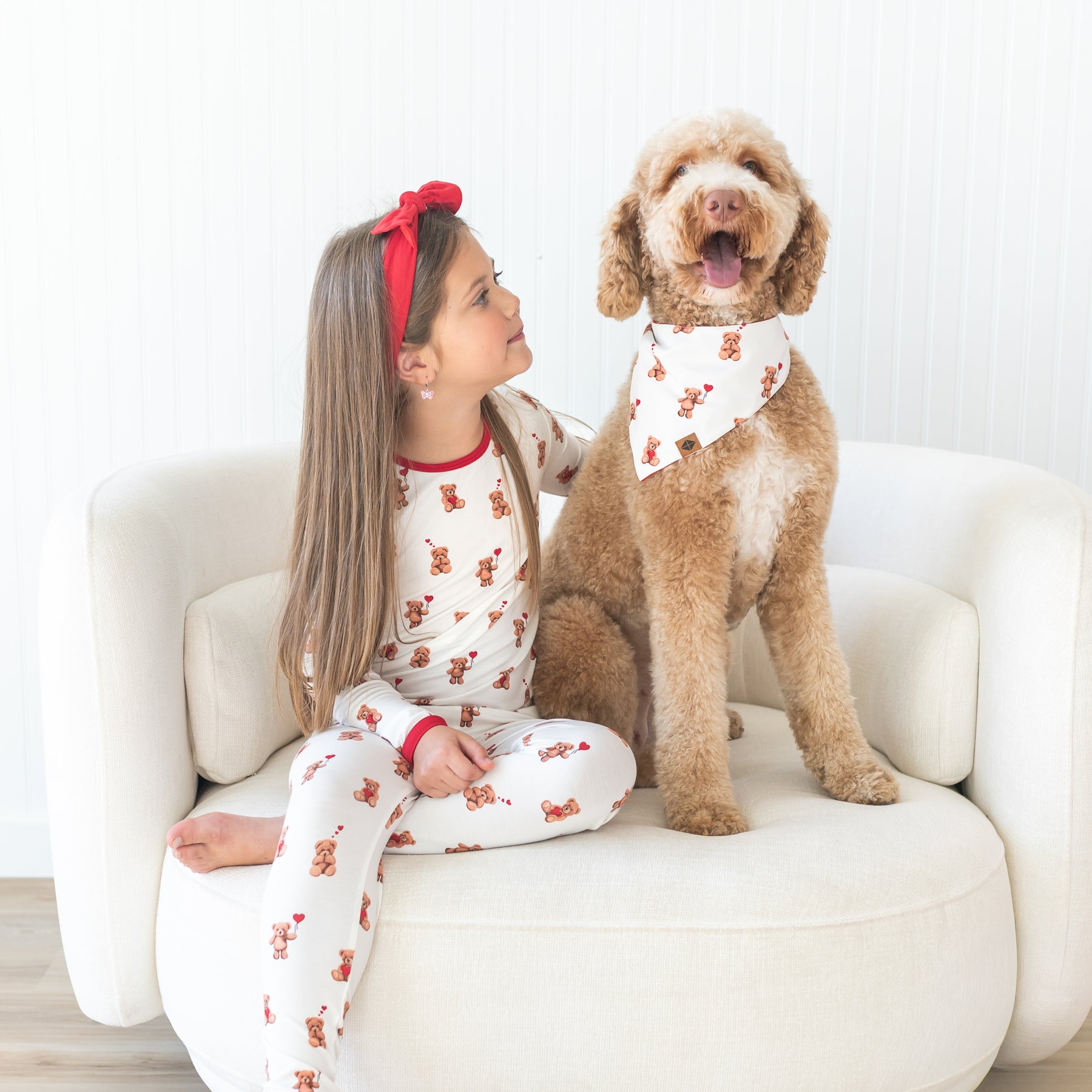 Young girl sitting on a cream sofa wearing the toddler pajamas in bear hearts sitting beside her dog who is wearing the Dog Bandana in Bear Hearts
