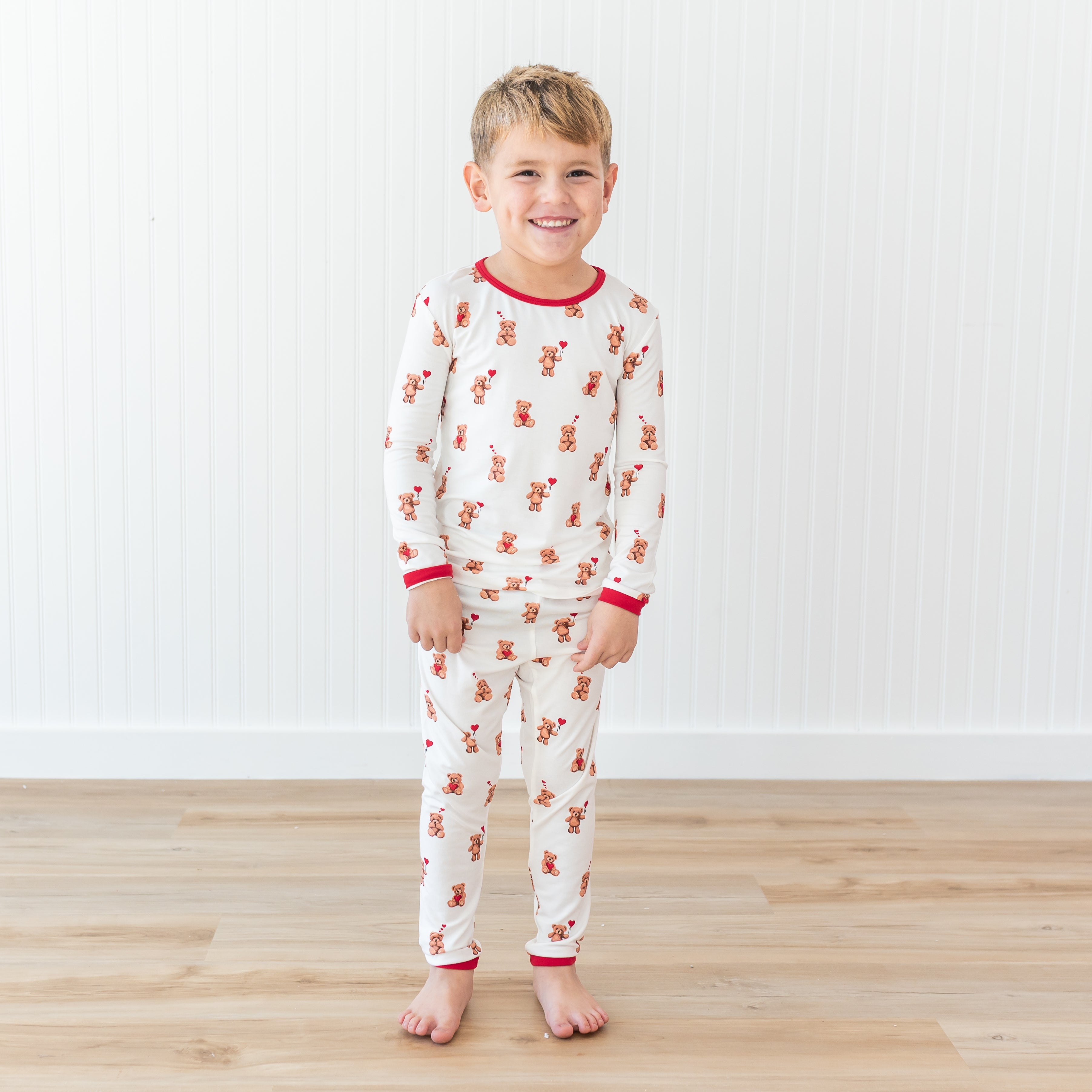 Smiling boy standing in front of a white paneled wall wearing the Long Sleeve Pajamas in Bear Hearts