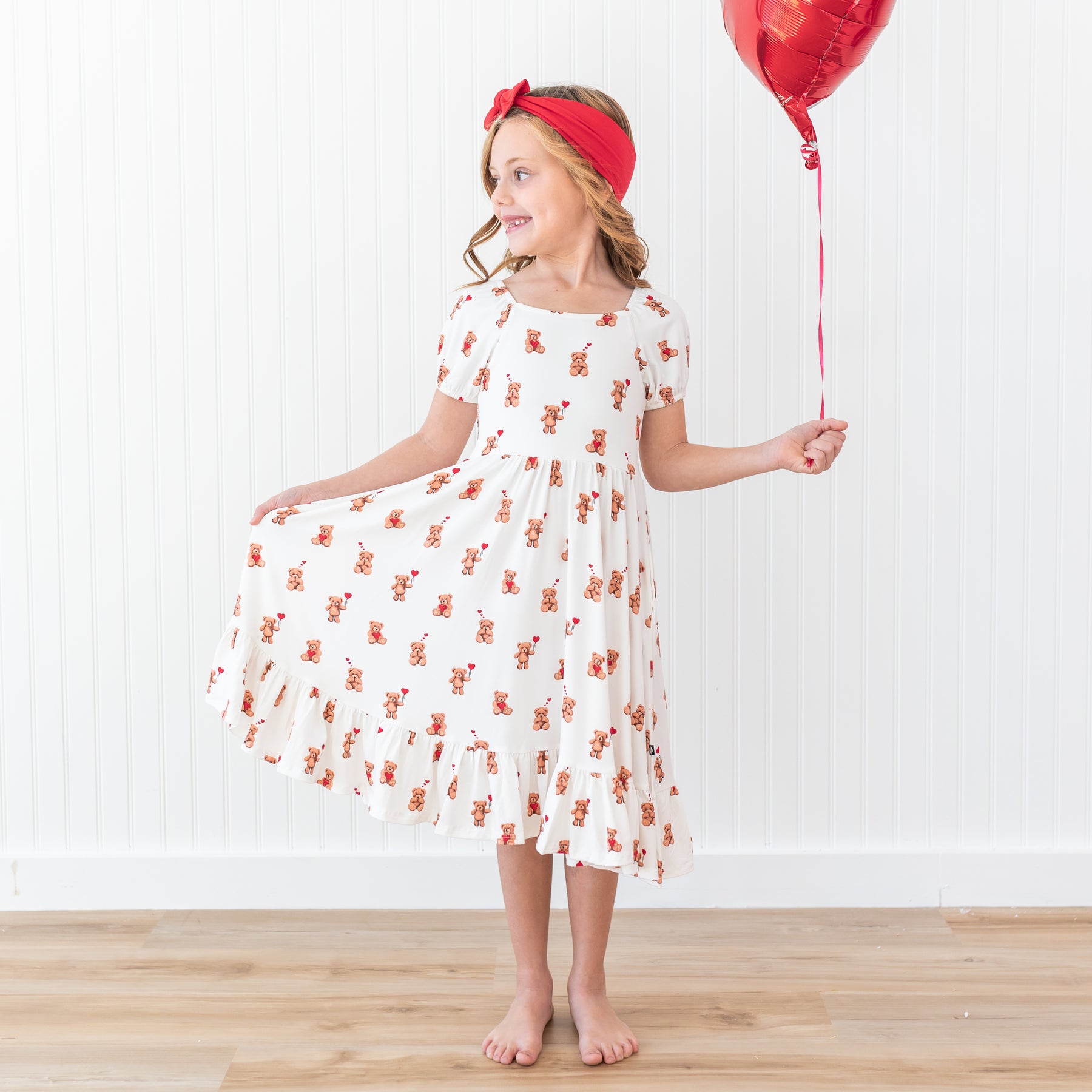 Young girl wearing the Puff Sleeve Twirl Dress in Bear Hearts with a Cardinal Knotted bow headband holding a red heart shaped balloon with one hand and the skirt of her dress in the other