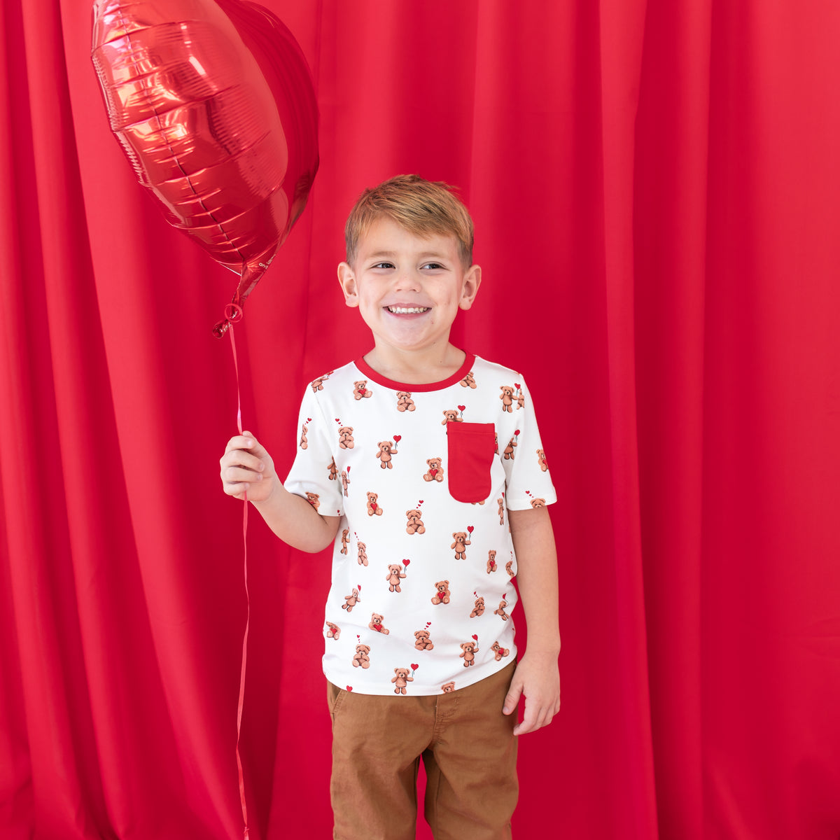 Smiling boy standing in front of a red curtain holding a red heart shaped balloon wearing the Toddler Crew Neck Tee in Bear Hearts