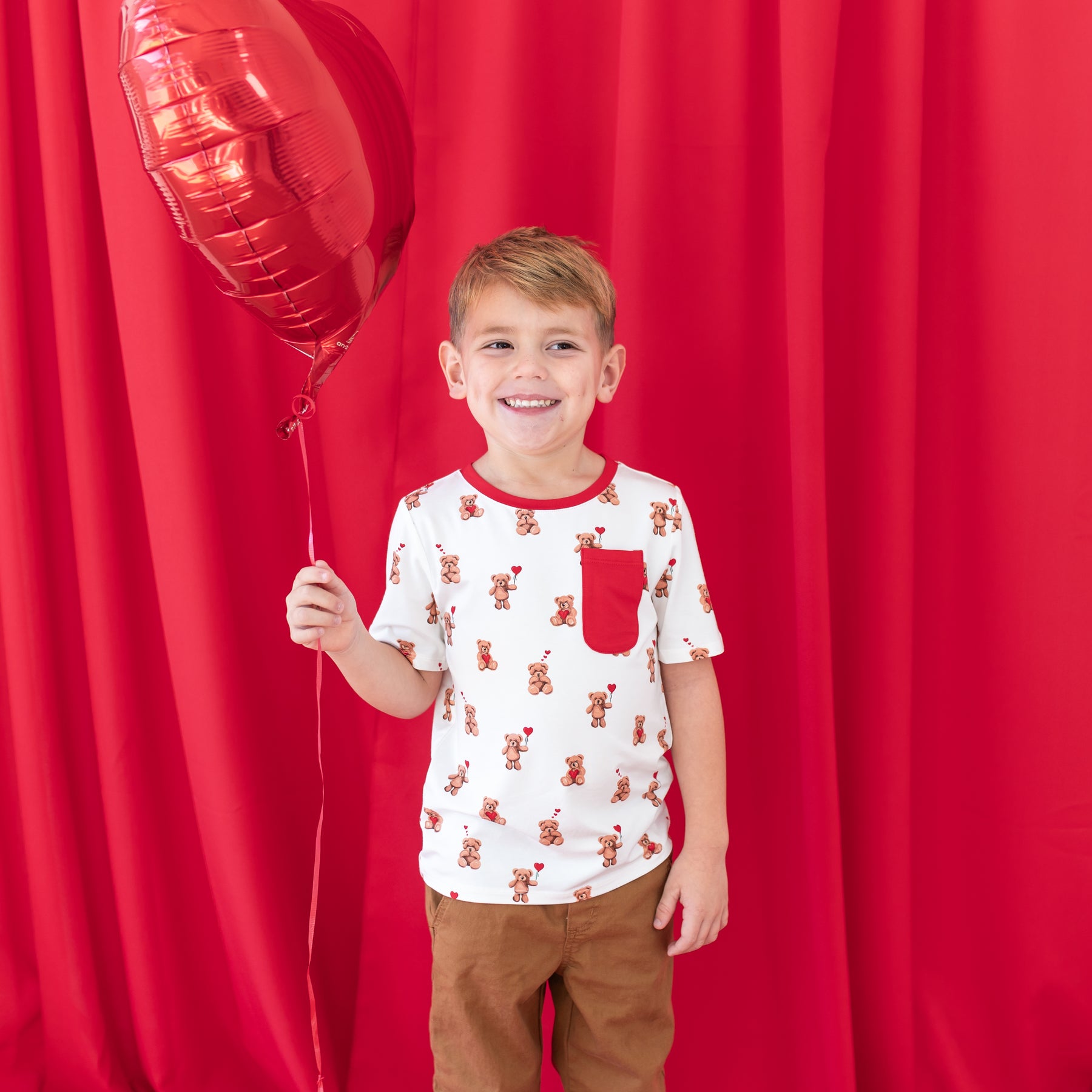 Smiling boy standing in front of a red curtain holding a red heart shaped balloon wearing the Toddler Crew Neck Tee in Bear Hearts