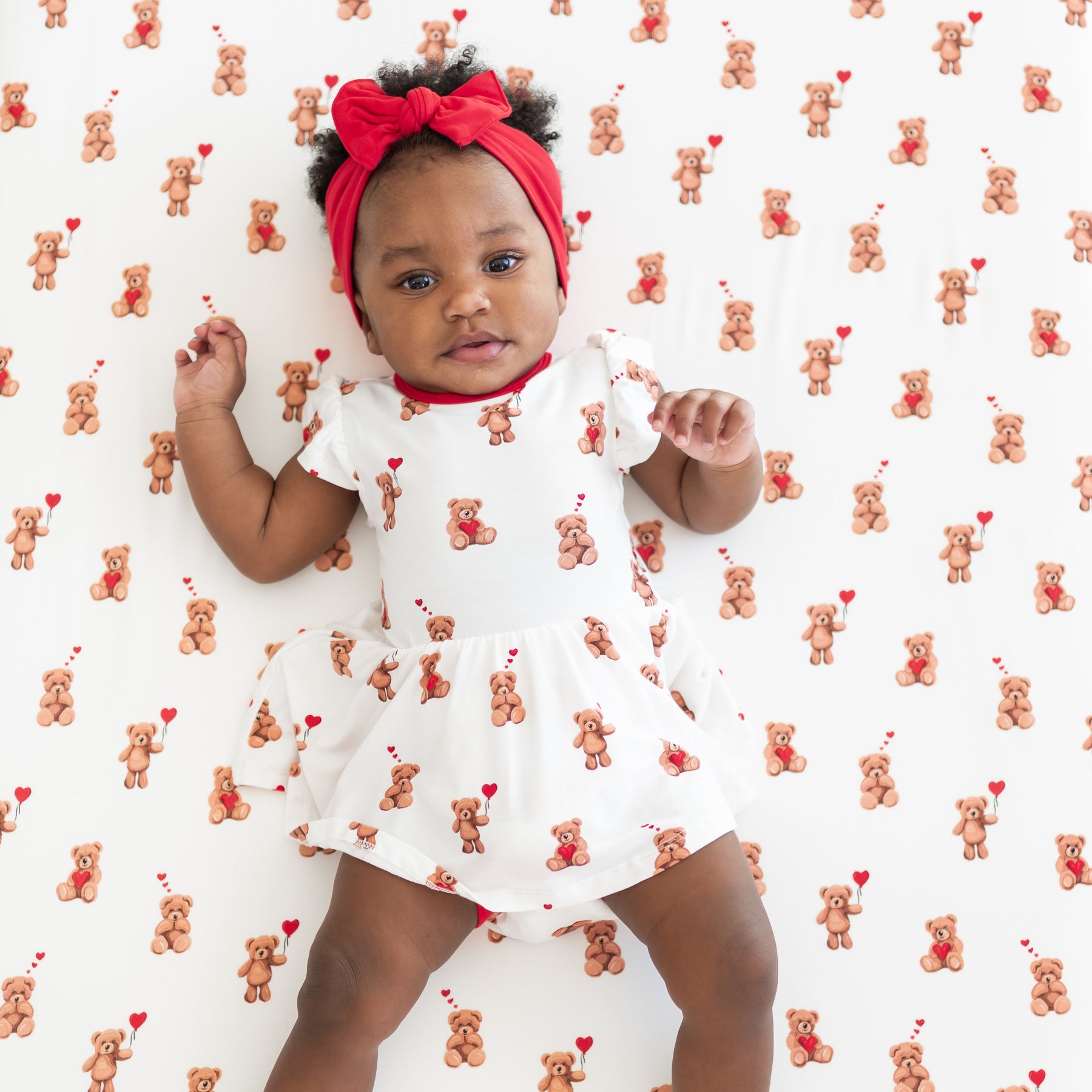 Toddler laying on a Bear heart crib sheet wearing the Twirl Bodysuit Dress in Bear Hearts paired with a Cardinal knotted bow headband