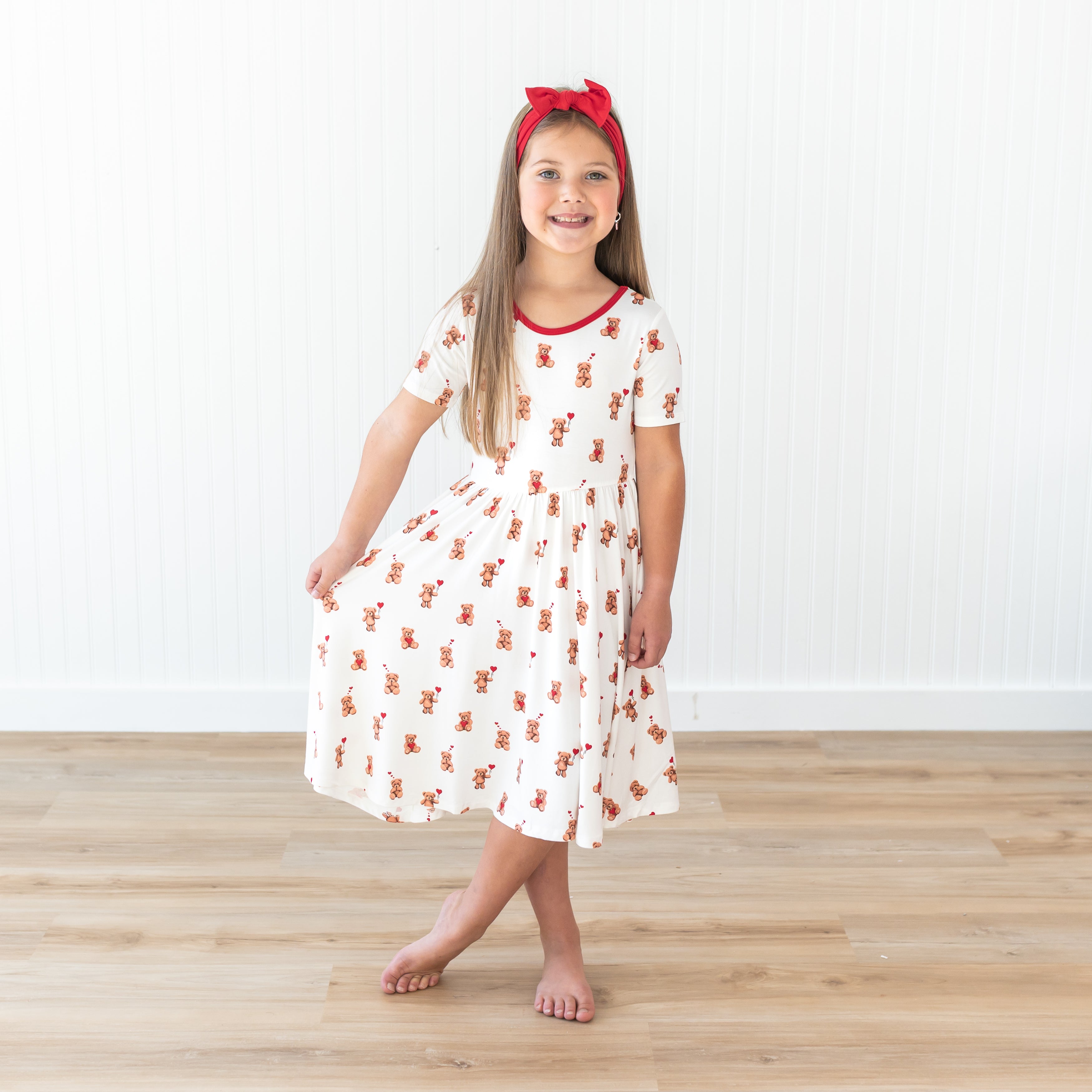 Young girl wearing the soft and flowy Twirl Dress in Bear Hearts paired with a Cardinal Knotted Bow standing in front of a white paneled wall