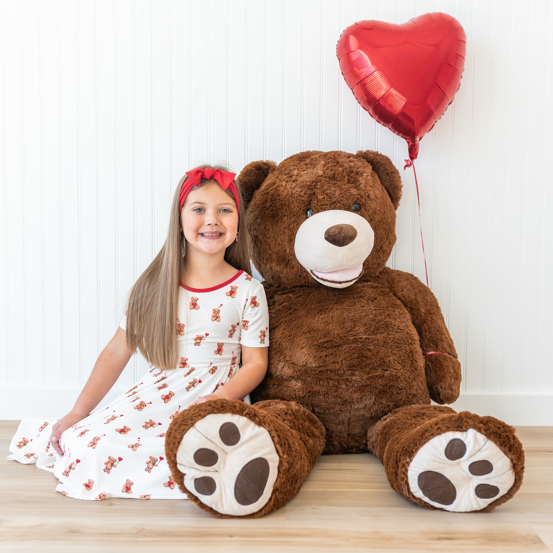 Young girl wearing the Twirl Dress in Bear Hearts sitting on the floor beside a large stuffed bear with a red heard balloon tied to its arm