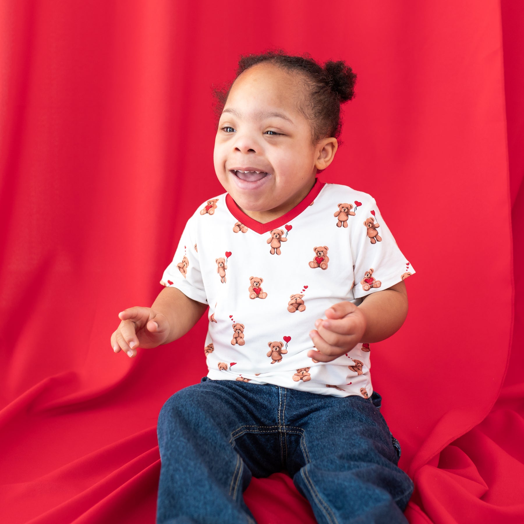 Young girl sitting in front of a red backdrop wearing the Toddler V-Neck Tee in Bear Hearts paired with medium wash colored jeans