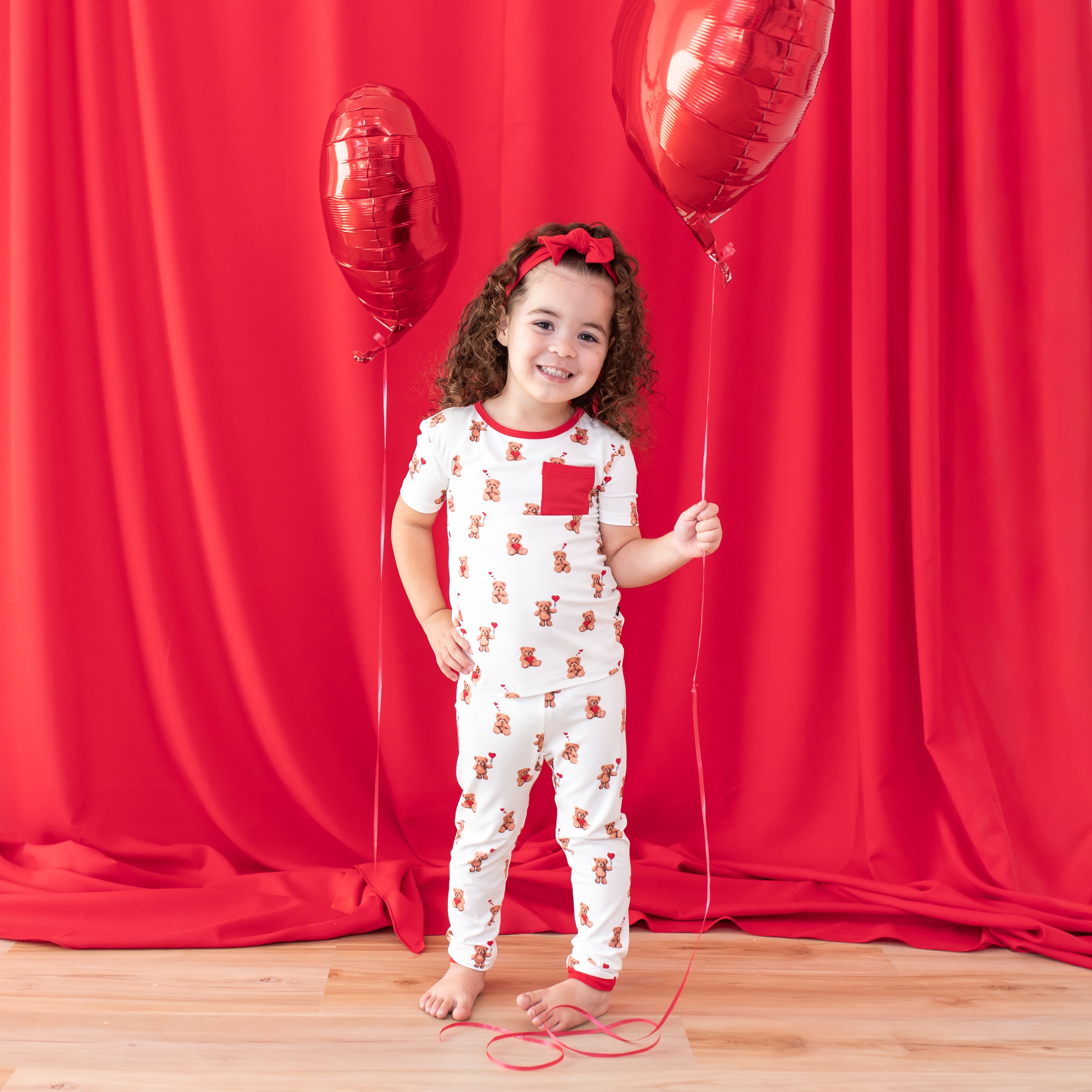 Young girl posing in the Short Sleeve with Pants Pajamas in Bear Hearts standing in front of a red curtain holding a red bear heart balloon